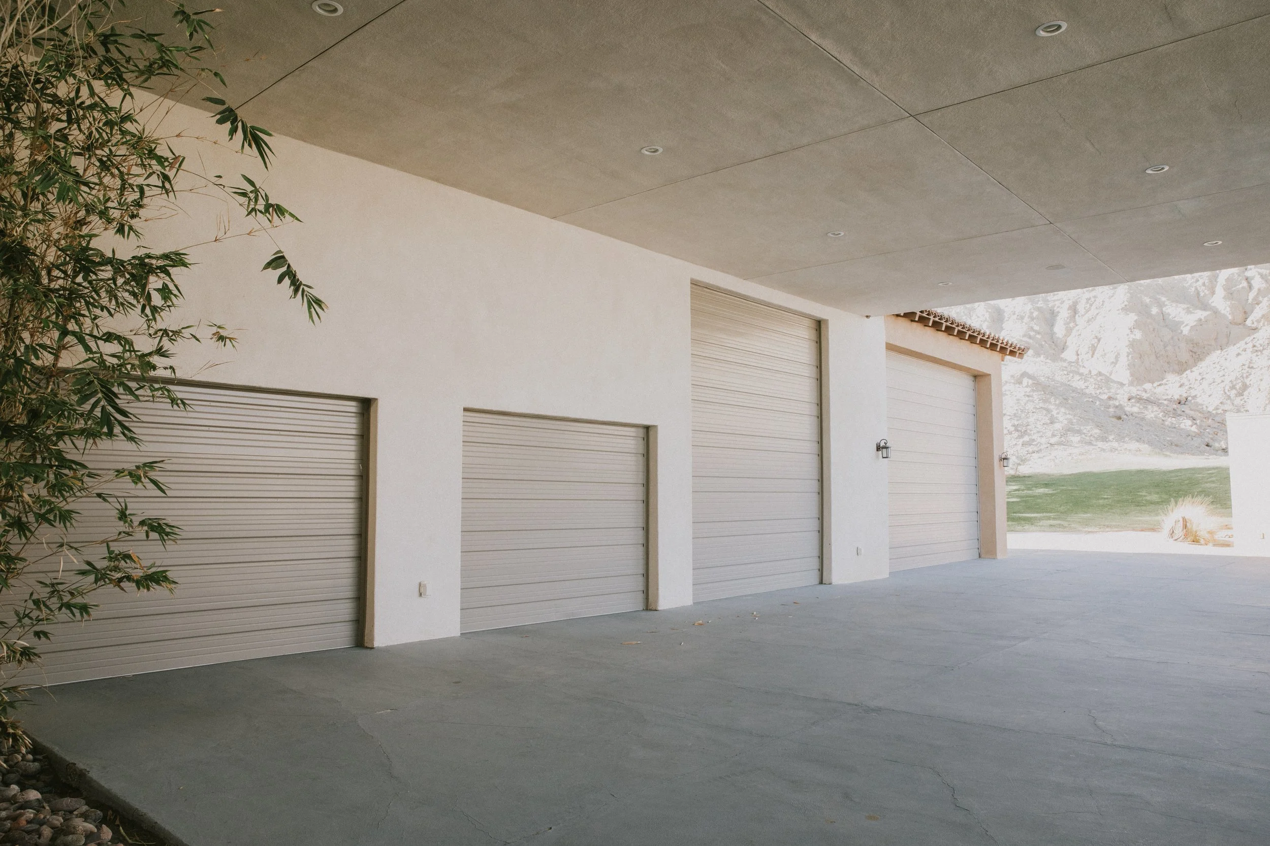 A photo of a garage with four closed cream-colored roll-up doors, a concrete driveway, beige walls, and a overhanging roof, with a mountainous landscape in the background.