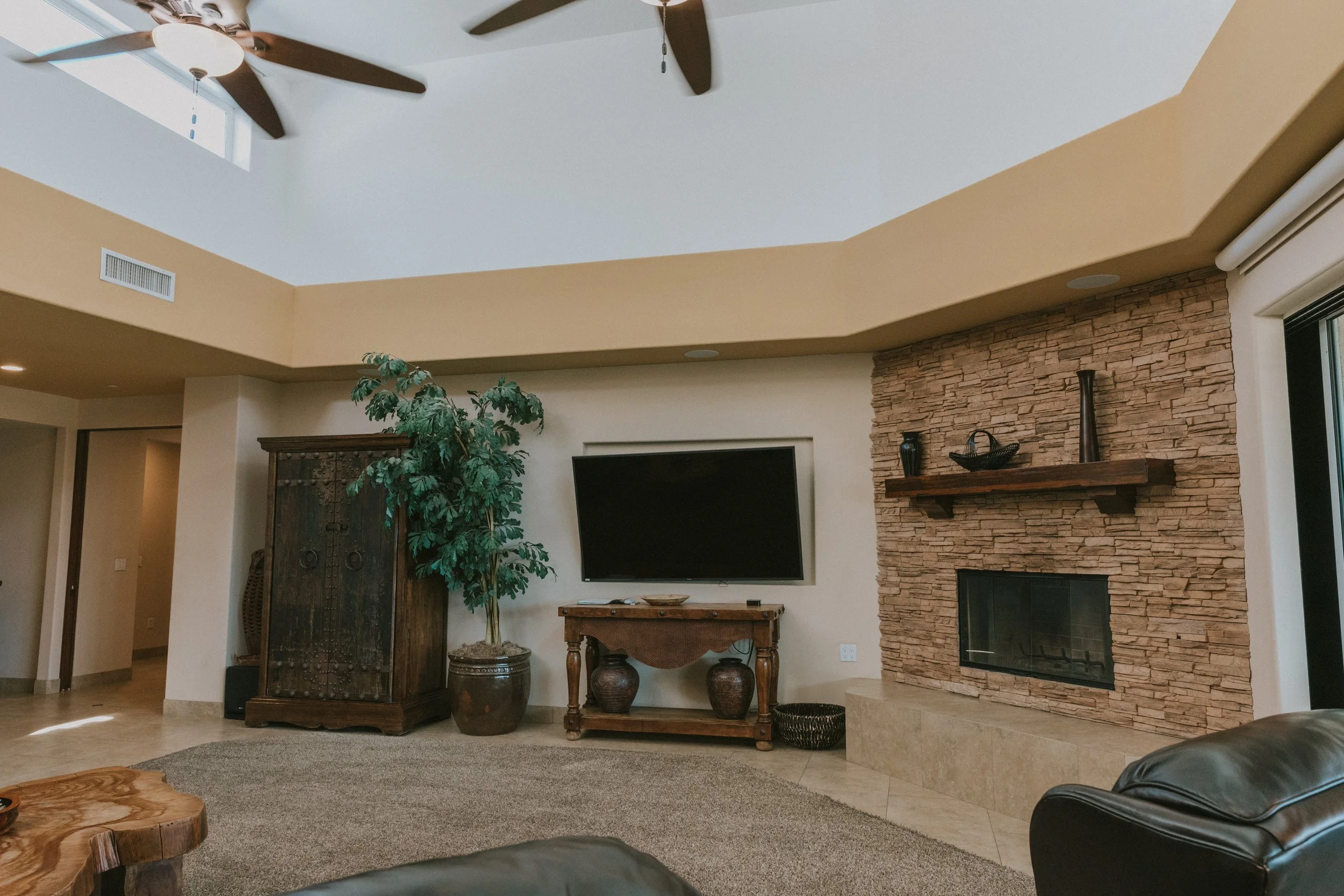 Living room with a stone fireplace, a mounted flat-screen TV, a wooden cabinet, and decorative vases. A large potted plant is near the furniture, and ceiling fans are visible on the ceiling.