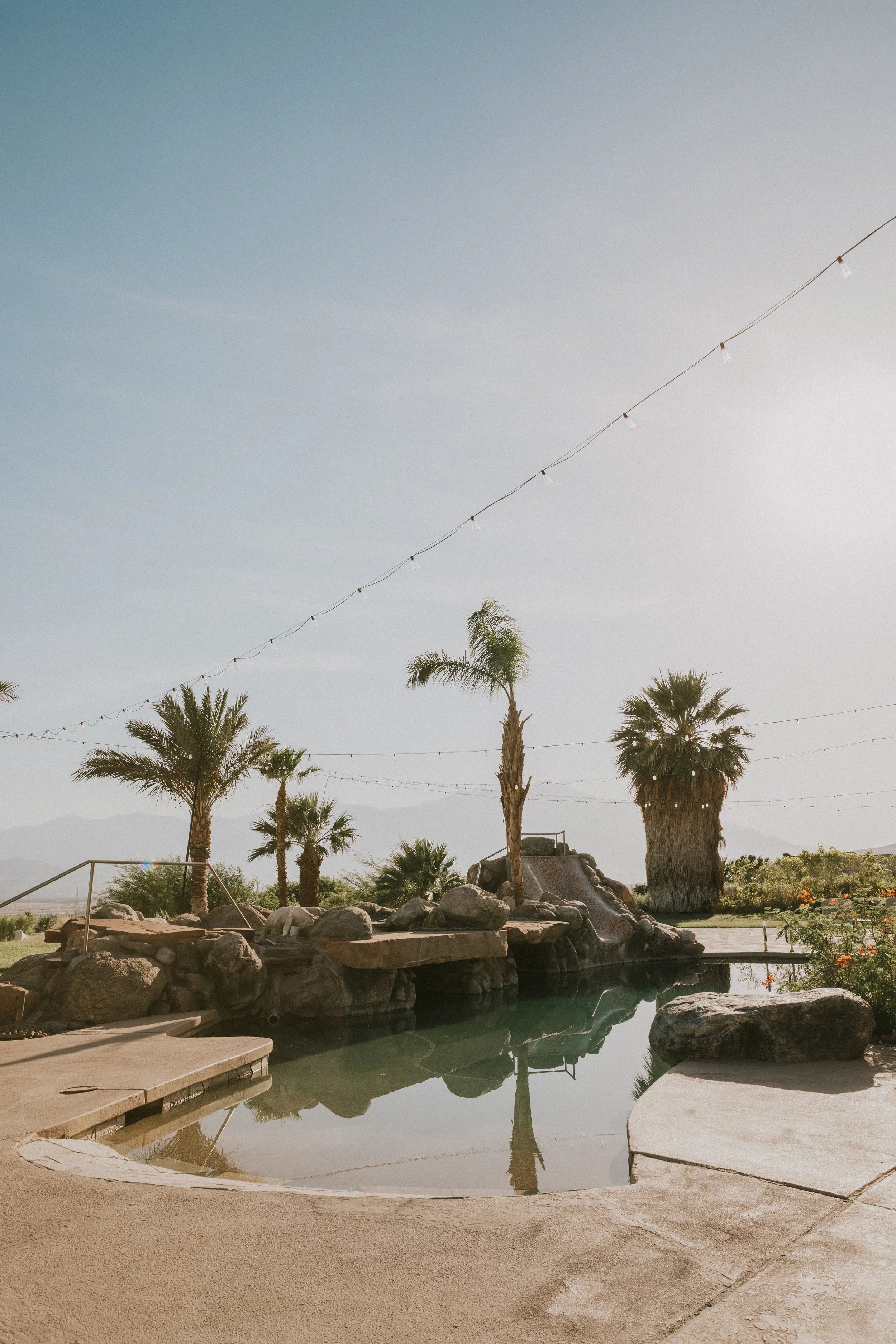 A serene outdoor landscape with a swimming pool, surrounded by rocks, palm trees, and a clear sky. String lights are strung across the scene, with mountains visible in the distance.