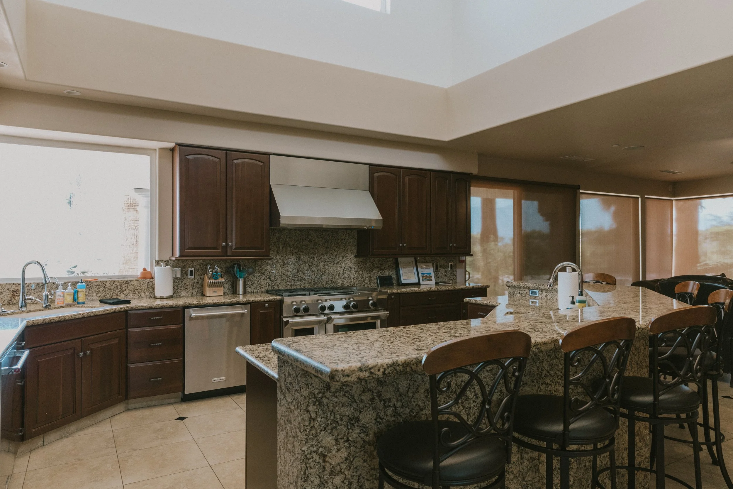 Kitchen with granite countertops, dark wooden cabinets, stainless steel appliances, and barstools around a large island. Windows with blinds let in natural light.