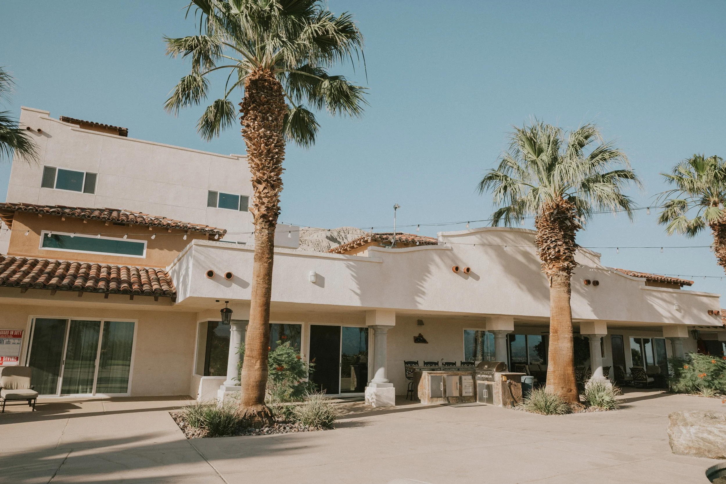 A residential building with white walls, tiled roof, and large sliding glass doors, surrounded by tall palm trees and outdoor seating, under a clear blue sky.