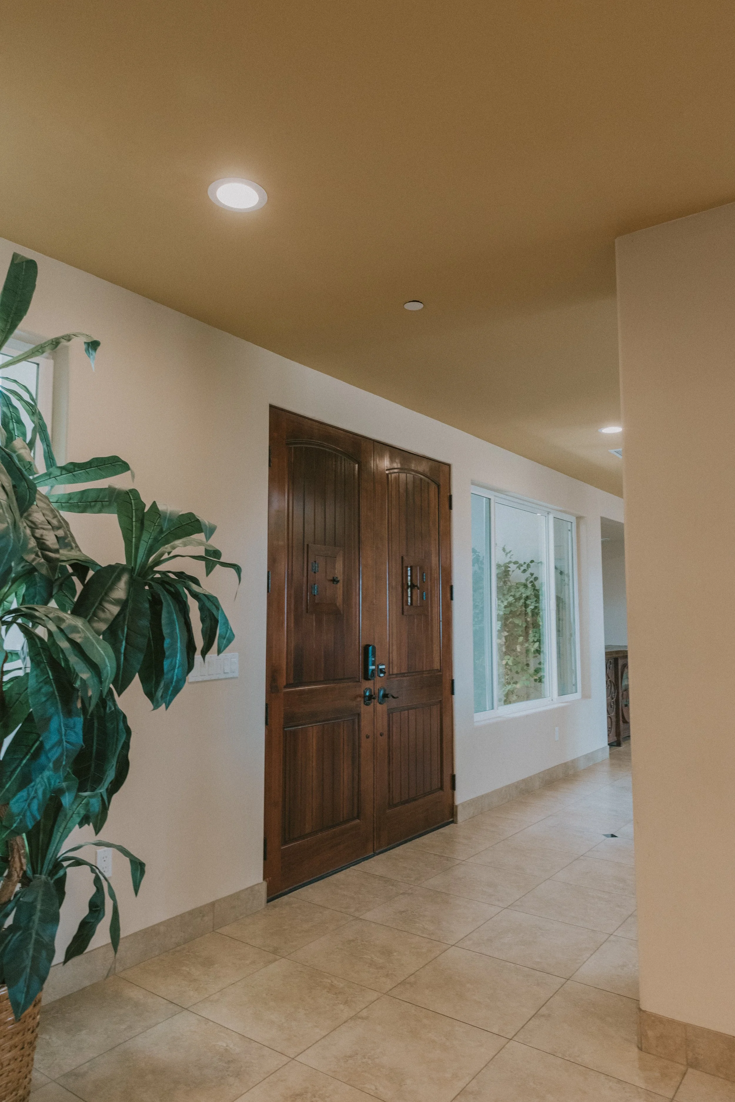 Interior view of a home with a wooden front door, a large window, tiled flooring, a potted plant, and recessed ceiling lights.