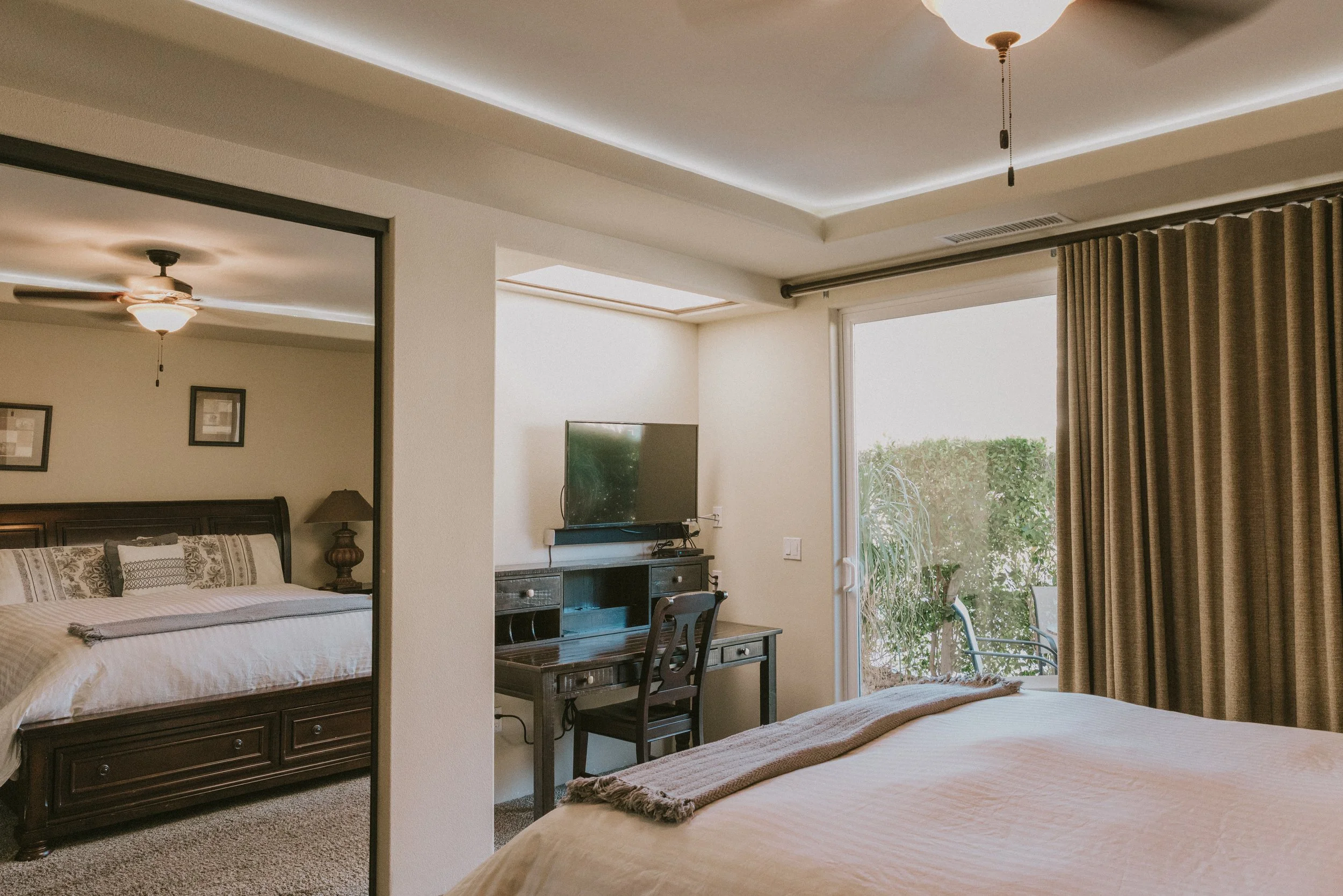 View of a bedroom with a bed, a dresser with a TV, and a small desk with a chair, adjacent to a sliding glass door with curtains that opens to a patio and greenery outside.