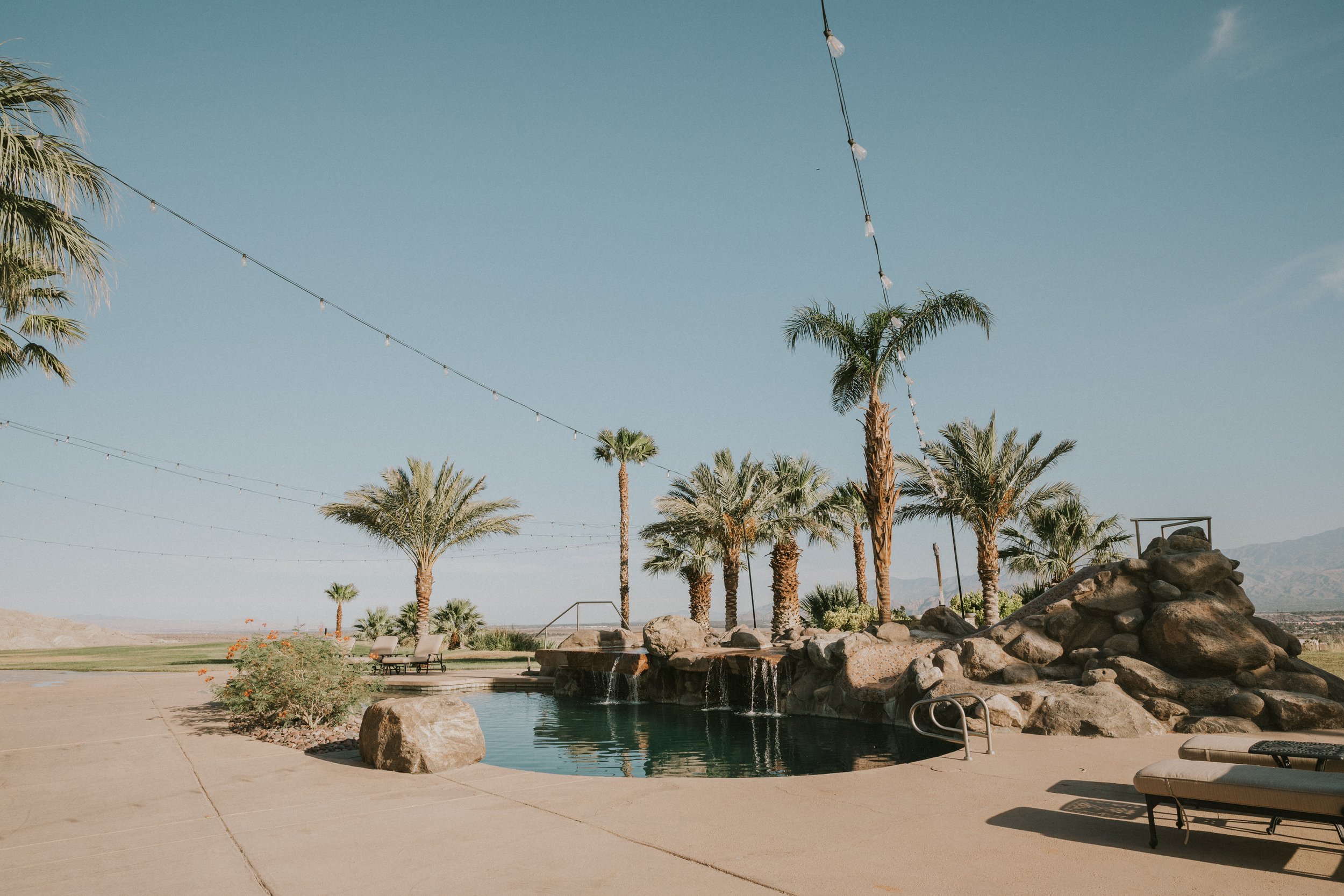 A swimming pool surrounded by palm trees, rocks, and lounge chairs in a desert landscape under a clear blue sky with string lights hanging overhead.