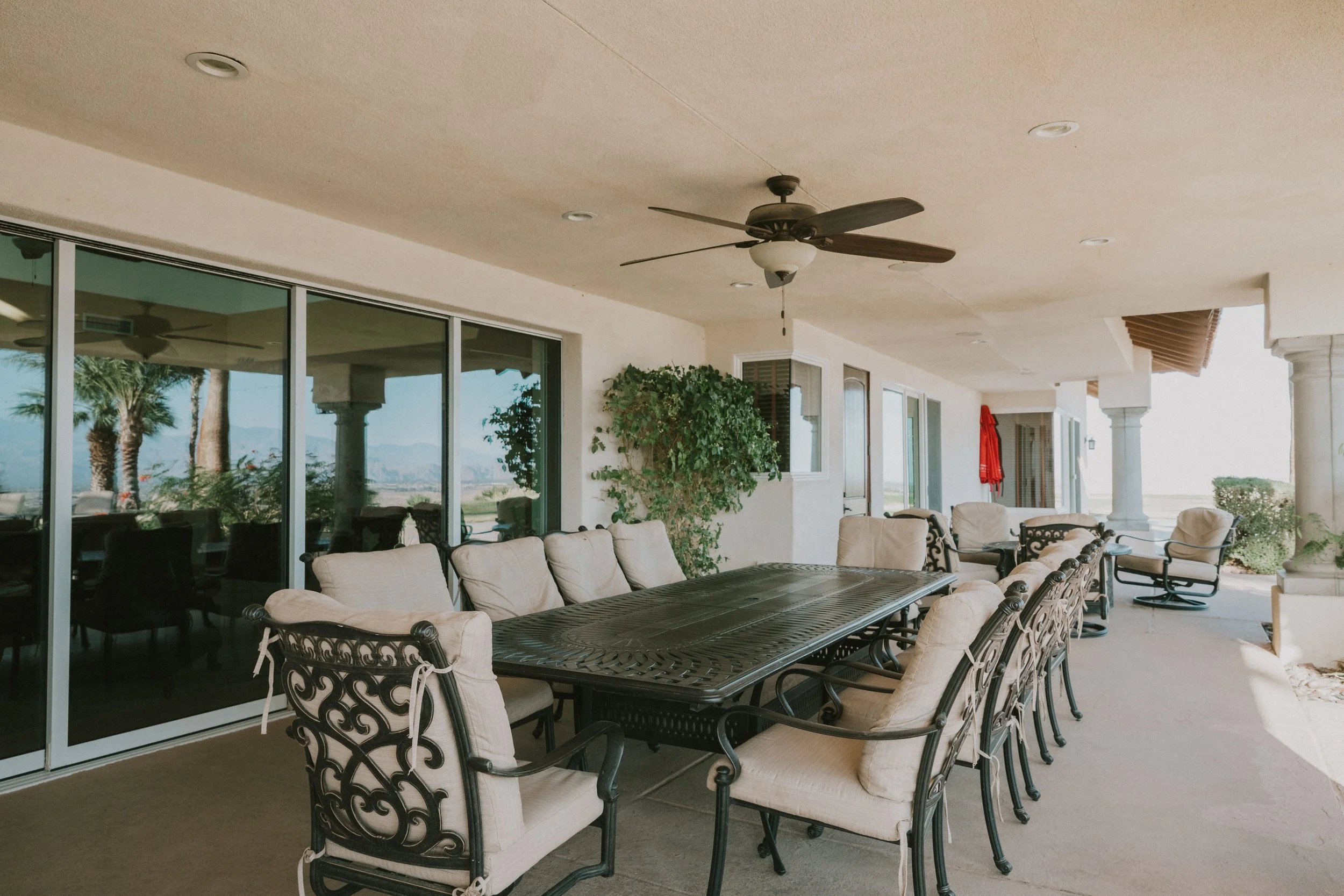 Covered outdoor patio area with a large dining table, cushioned chairs, ceiling fan, potted plants, and large glass sliding doors leading inside. Palm trees and mountains are visible in the background.