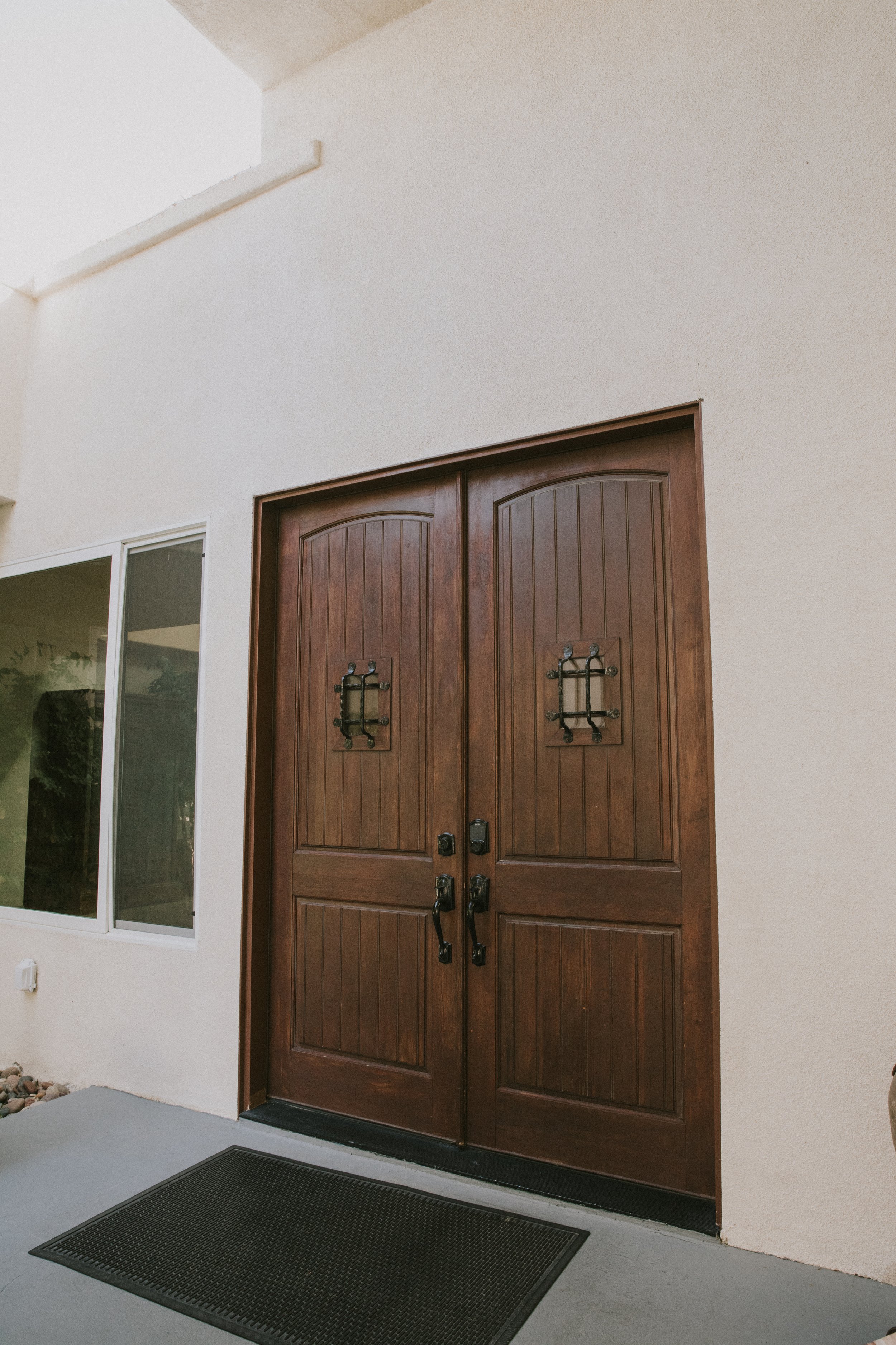 A wooden double front door with black handles and decorative hardware, set into a beige stucco wall with a small window or sliding glass door to the left and a black outdoor rug on the concrete porch.