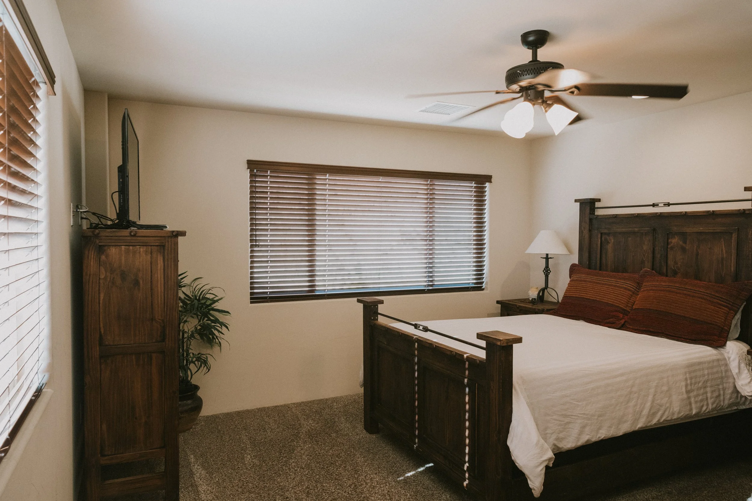 A bedroom with a wooden bed, brown pillows, a nightstand, a lamp, a window with brown blinds, a ceiling fan, and a TV on a wooden cabinet.