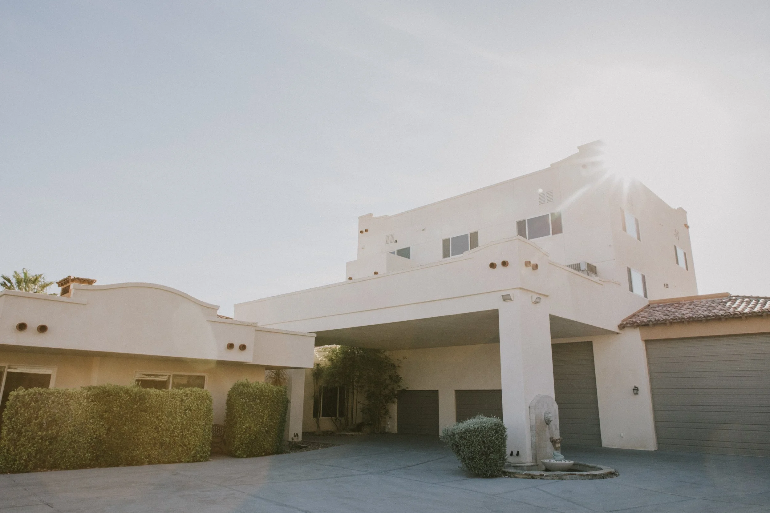 Modern white multi-story house with a curved balcony, large windows, and a garage, surrounded by bushes and trees, under a clear sky.