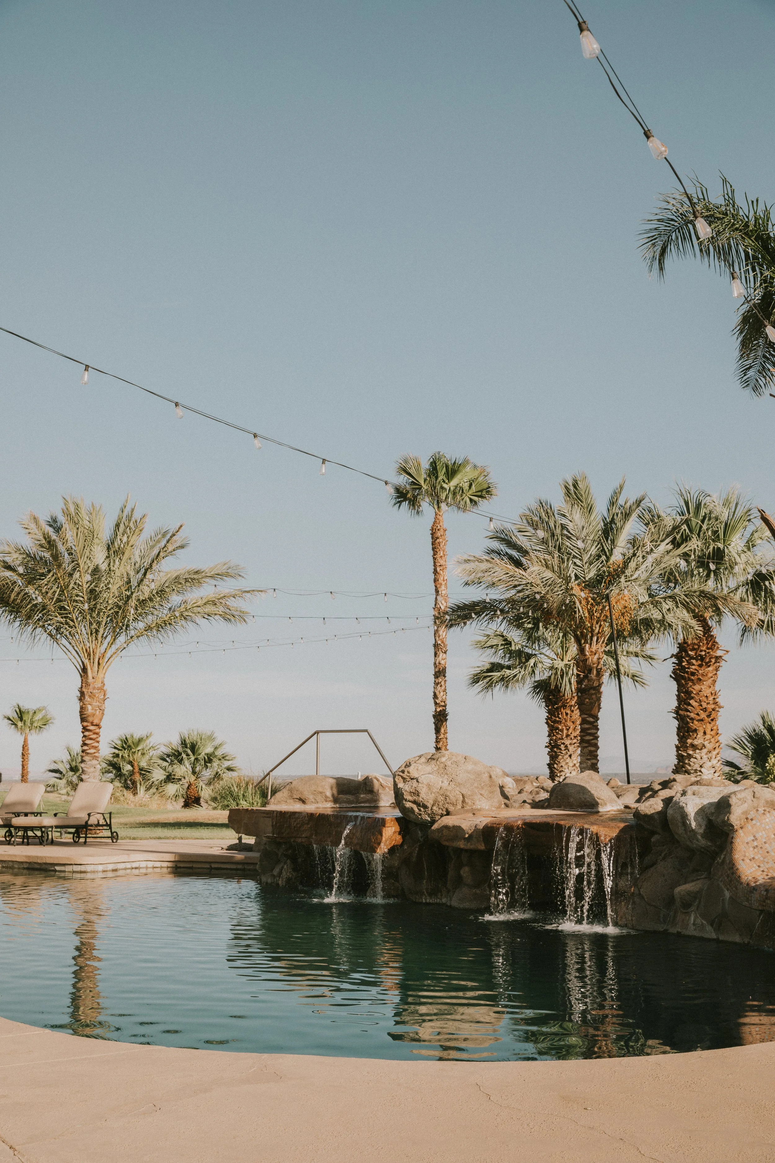 Pool area with a waterfall feature, surrounded by palm trees, outdoor lounge chairs, and string lights against a clear blue sky.