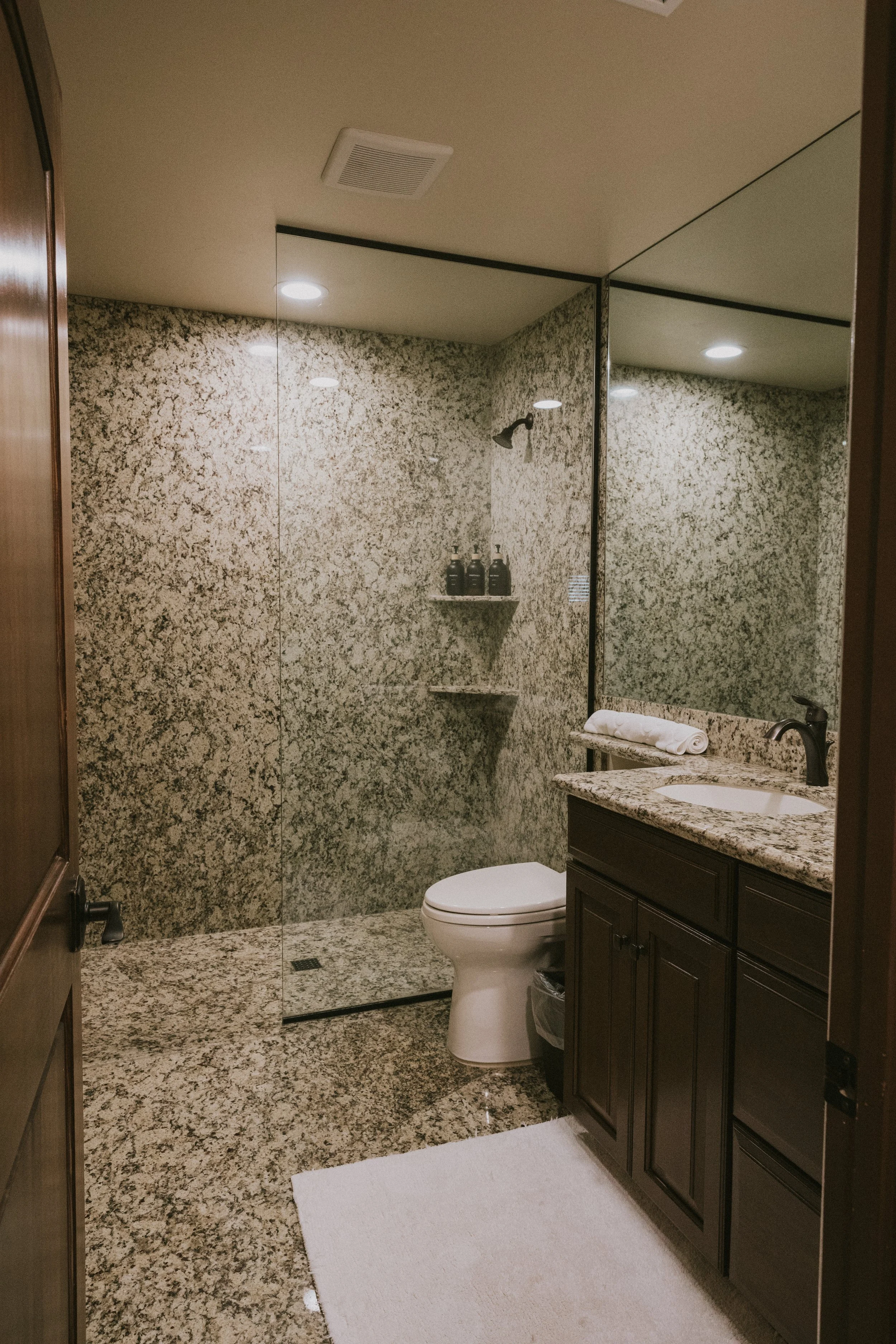 Bathroom with granite walls and floor, a white toilet, dark brown vanity with a granite countertop, a mirror with a black frame, a towel with a towel holder, soap dispenser, and a shower with glass door and mounted showerhead.
