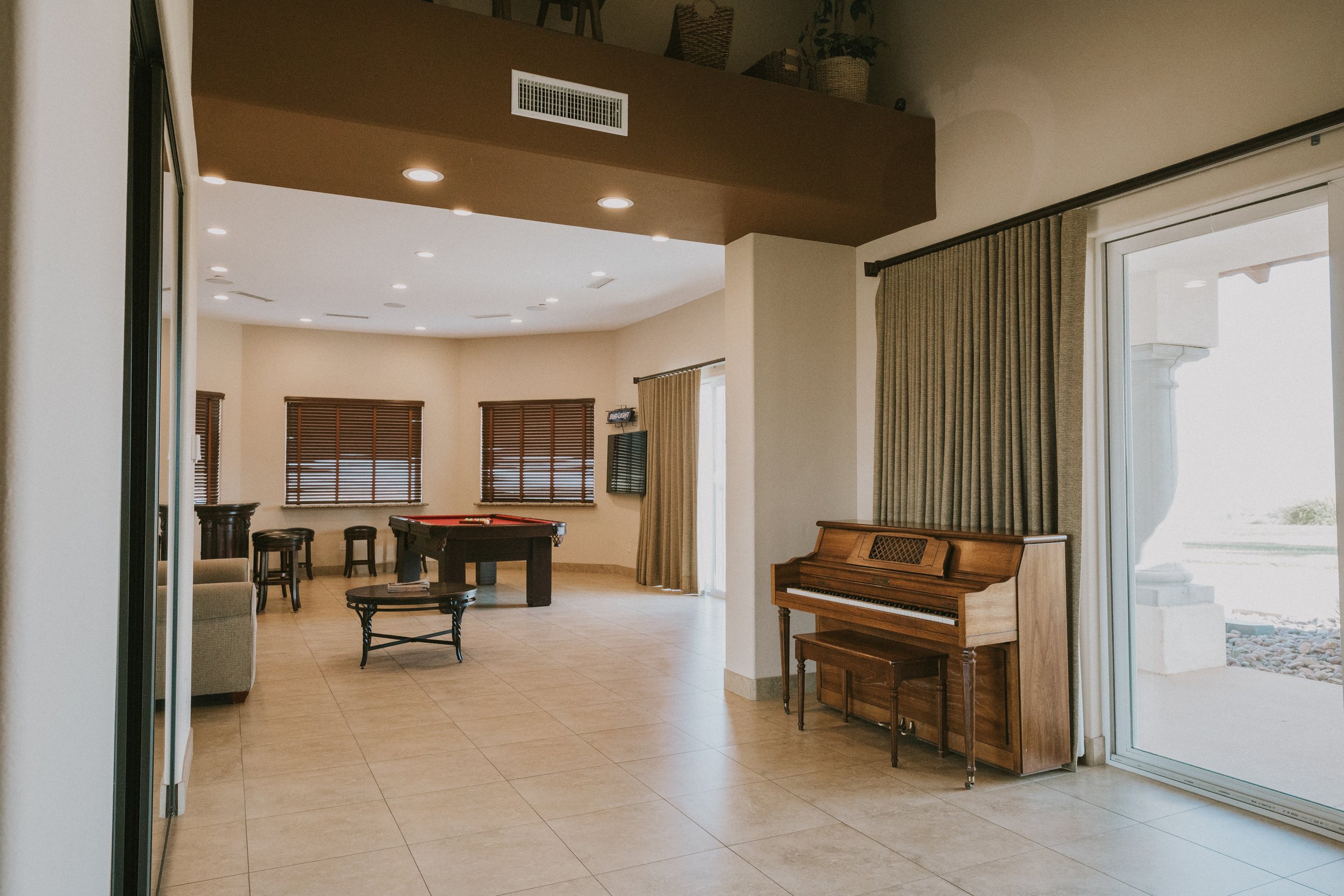 Interior view of a living area with a piano, a pool table, chairs, large windows with blinds, and sliding glass doors leading outside.