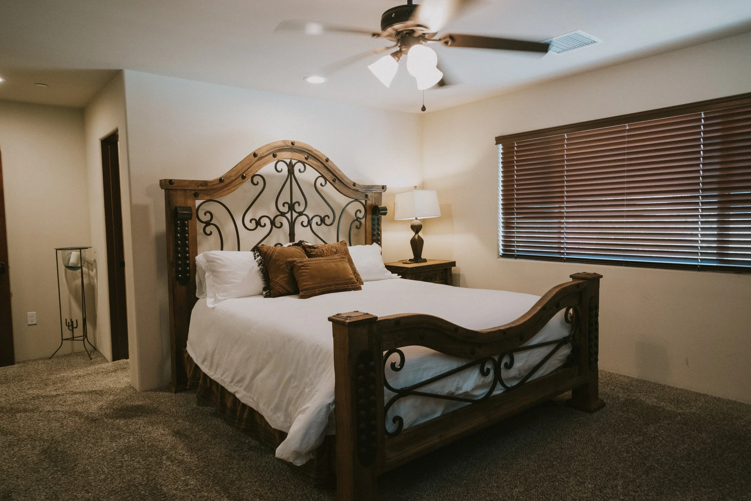 A bedroom with a rustic wooden bed frame featuring ornate metalwork, white bedding, brown decorative pillows, a wooden nightstand, a table lamp, a large window with wooden blinds, and a ceiling fan with light fixtures.