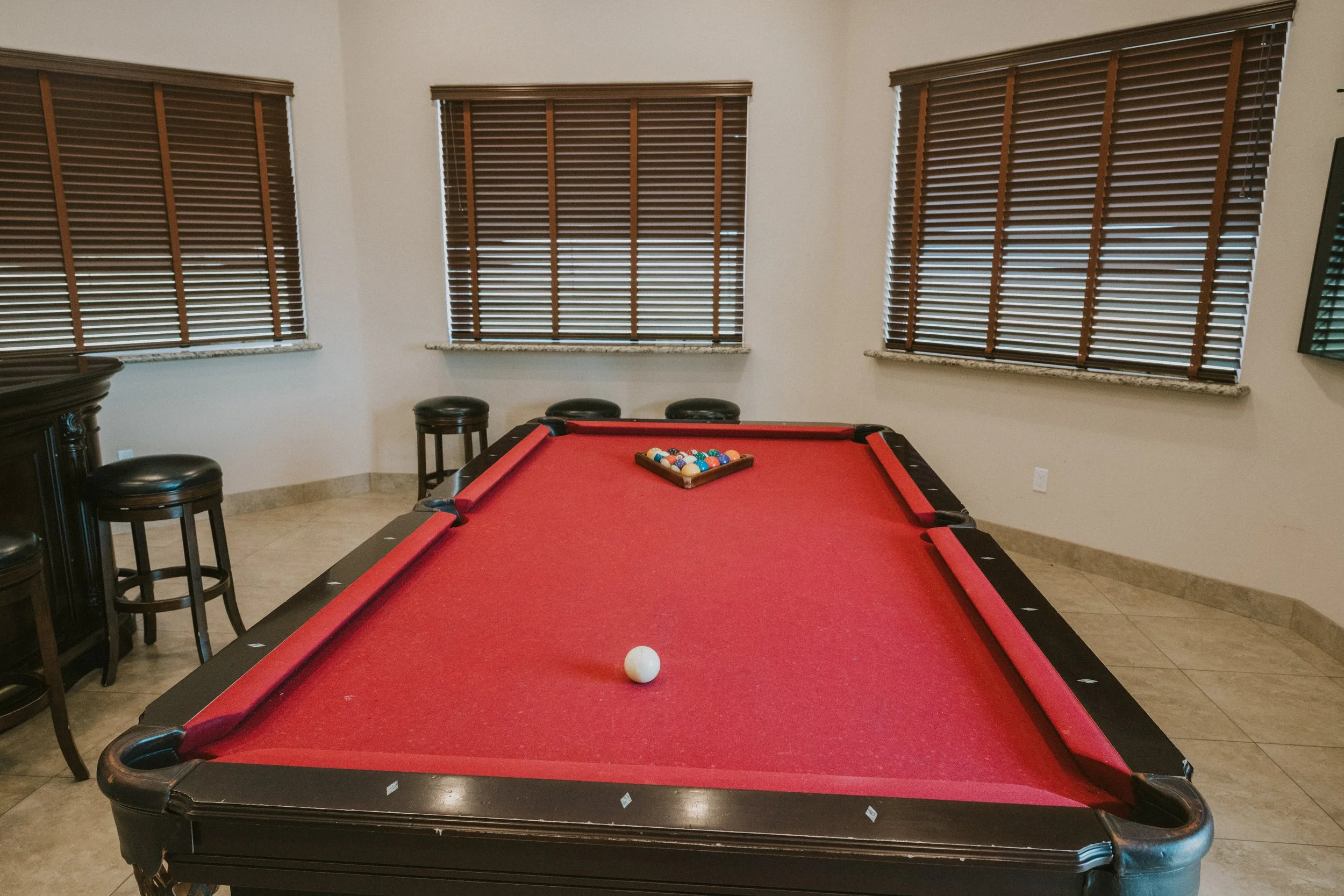 A red pool table with a white cue ball and a rack of billiard balls in the center, surrounded by three black stools, three large windows with closed wooden blinds, and a granite windowsill in a beige-walled room.
