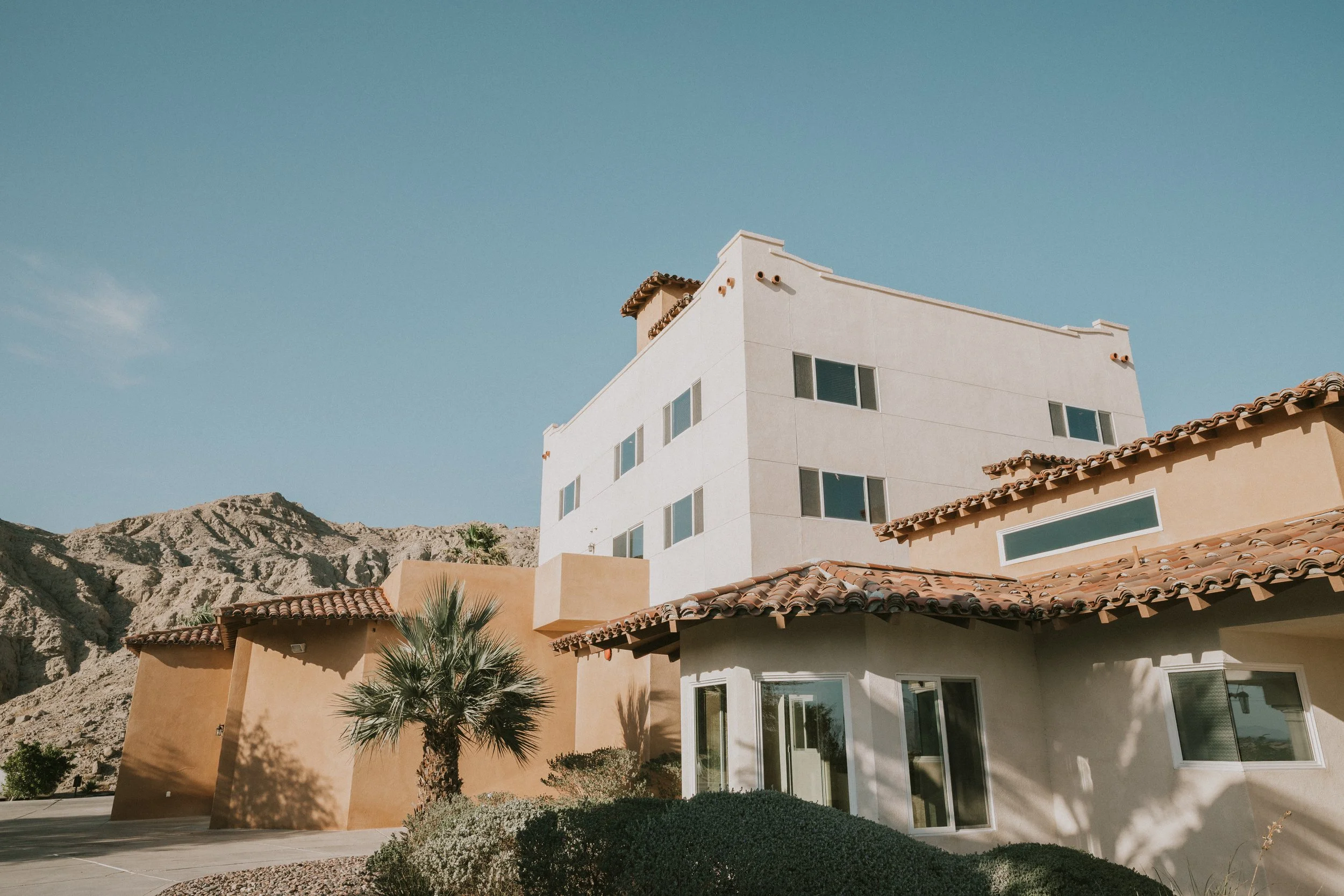 A modern multi-story house with a flat roof and large windows, situated in a desert landscape with mountains in the background. The house has a white exterior with some beige sections and a tiled roof on a smaller connected structure. There is a smal