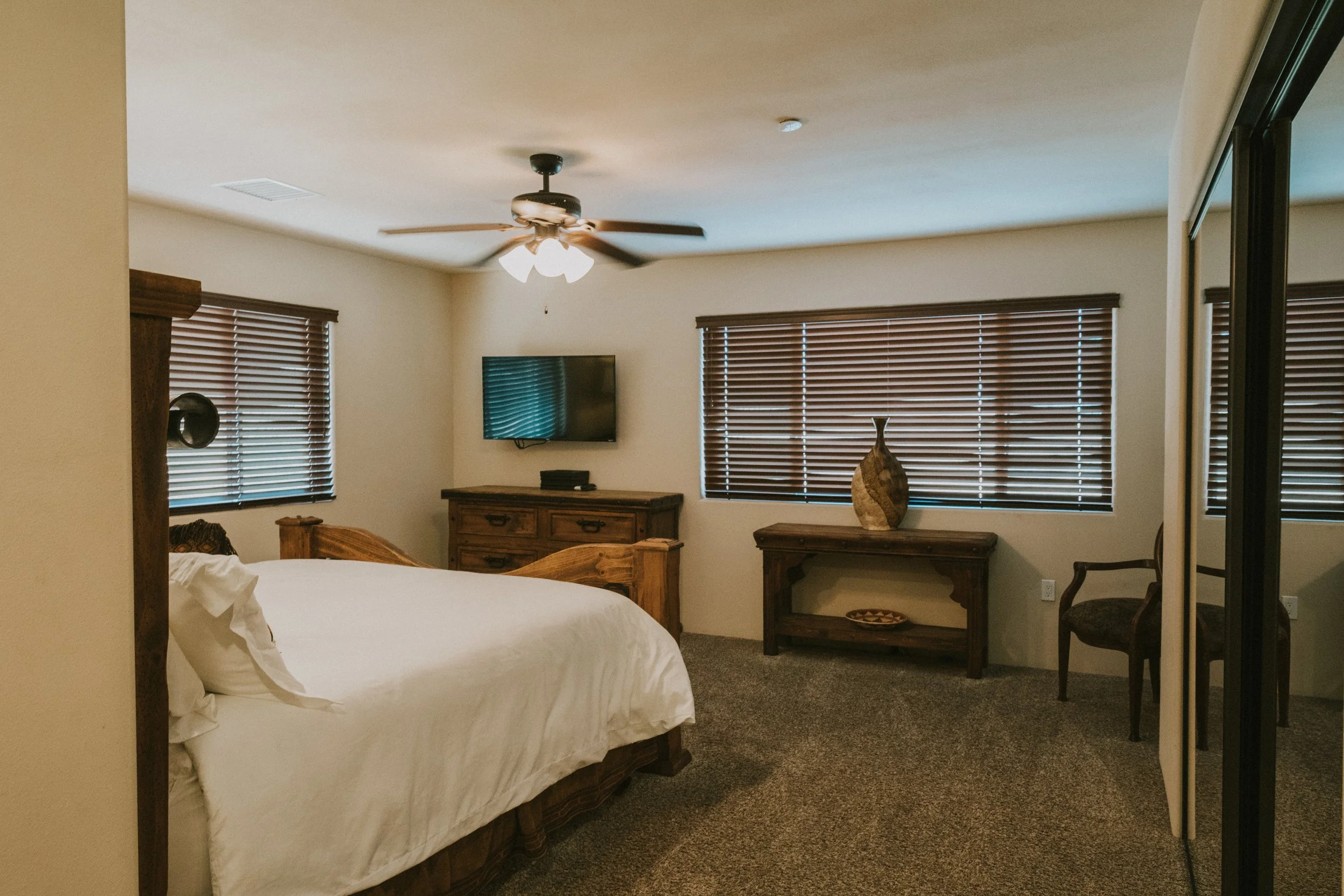 A bedroom with a wooden bed frame and white bedding, wooden furniture, a wall-mounted television, and blinds on the windows.