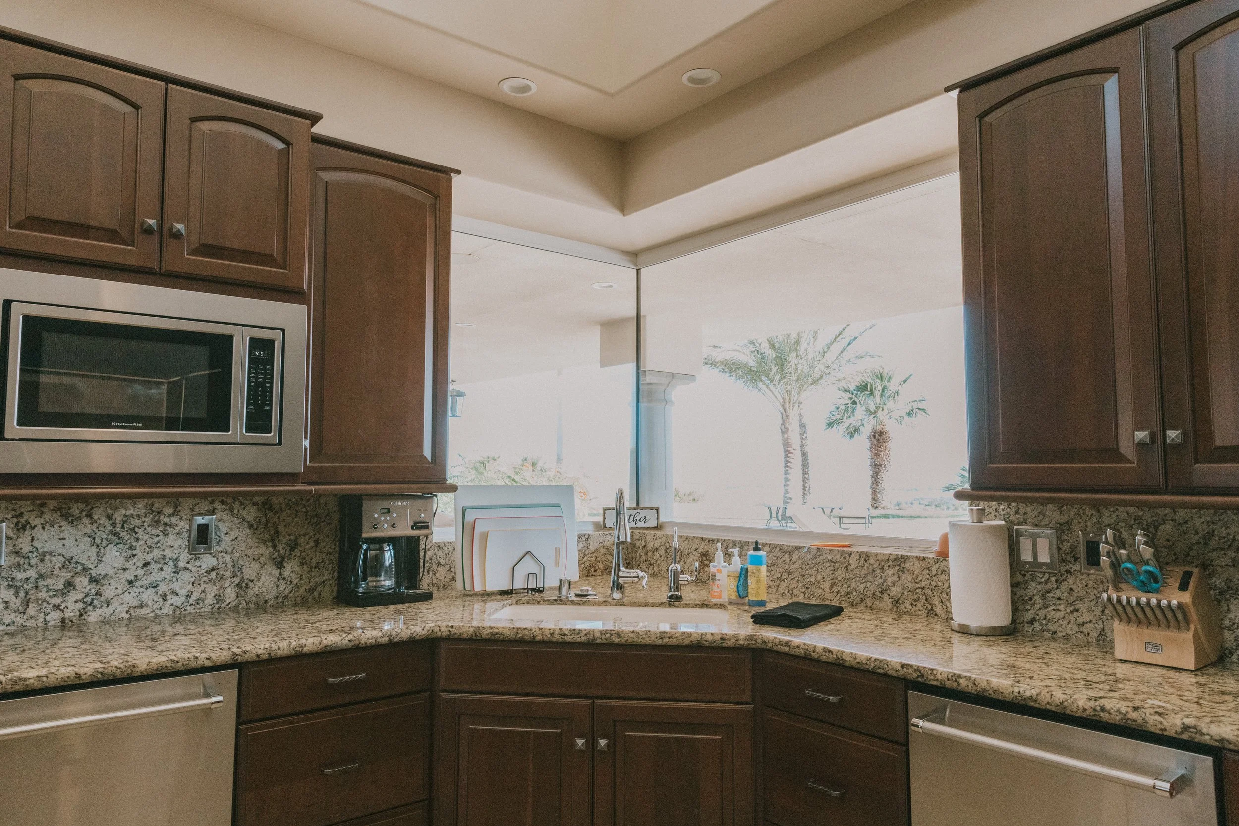 Kitchen with wooden cabinets, granite countertop, microwave oven, and a view of palm trees through the window.