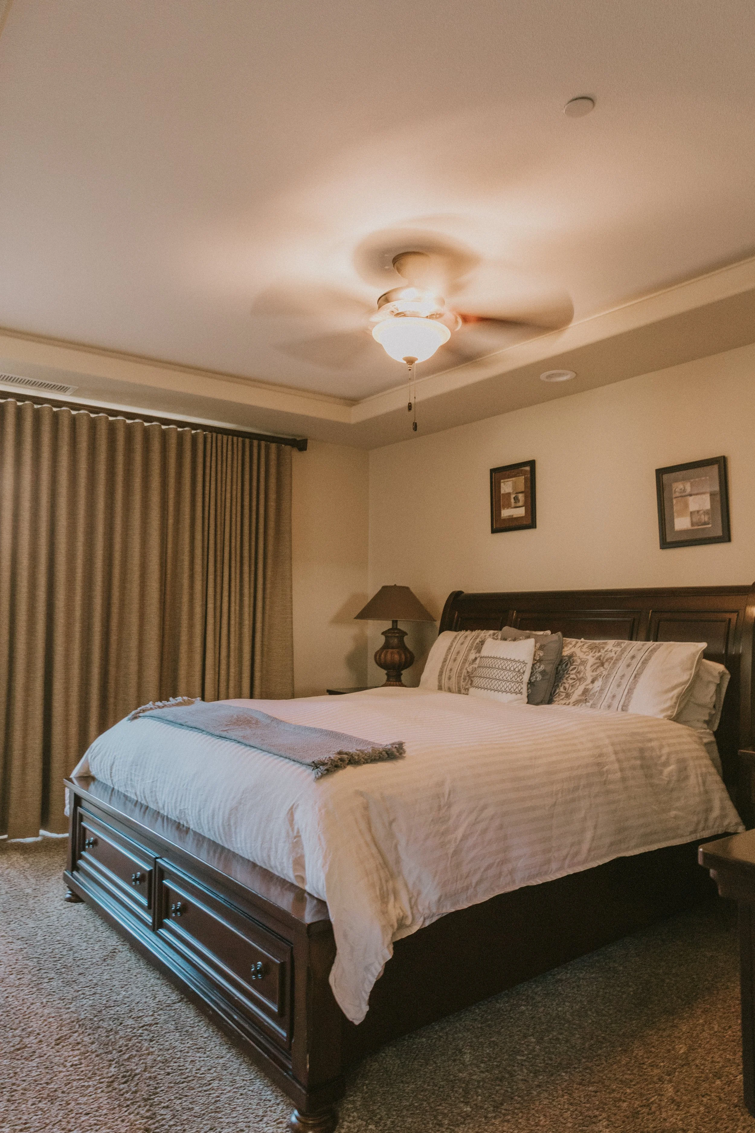 A neatly made bed with white and beige bedding, decorative pillows, a dark wooden headboard, and a brown blanket at the foot of the bed in a bedroom with beige carpet, beige walls, two framed pictures, curtains, and a bedside lamp.