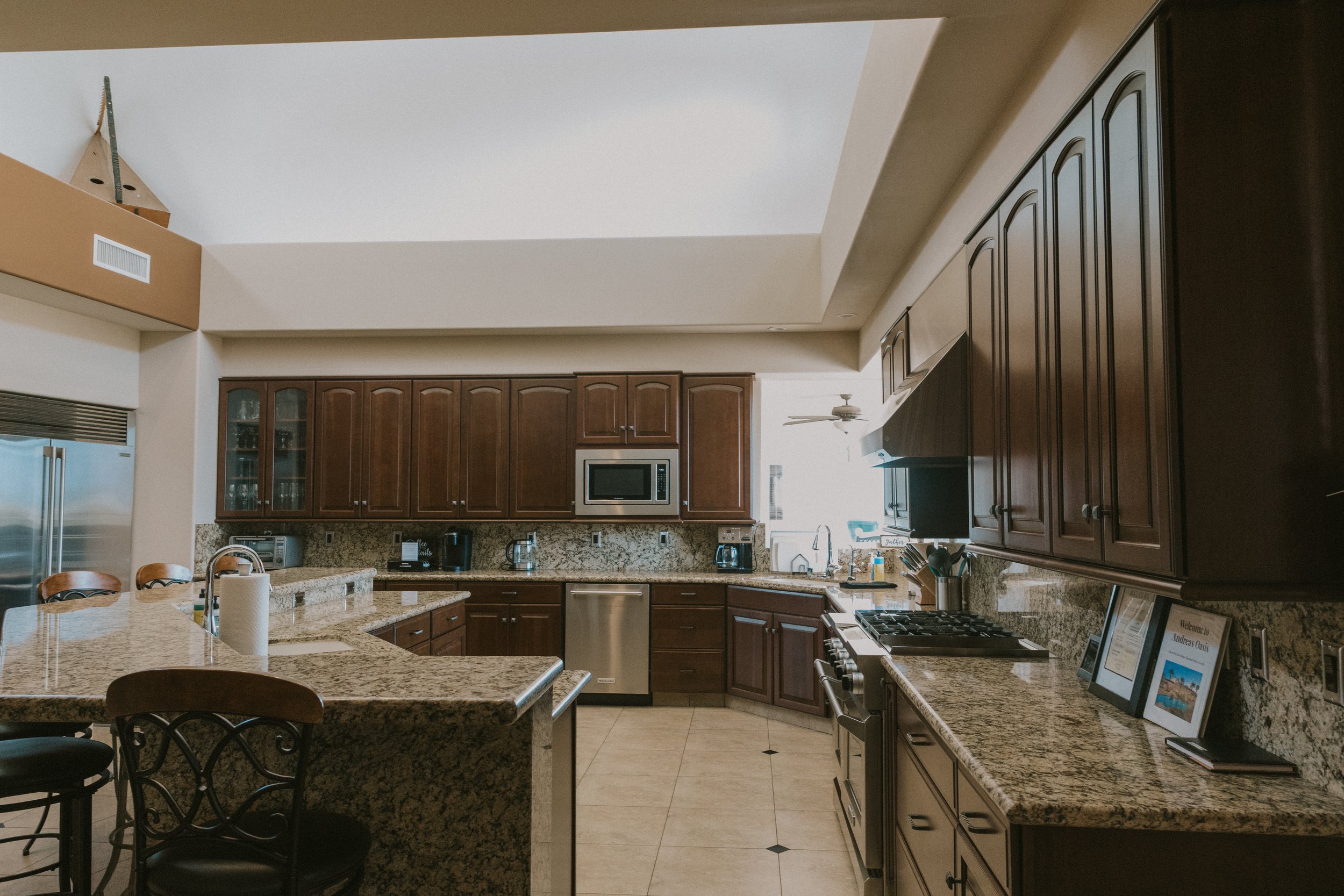 Kitchen with dark wooden cabinets, granite countertops, stainless steel appliances, and a large island with seats. There are small drawers and cabinets below the countertops and a microwave built into the cabinets. The ceiling has a fan and a vent, a