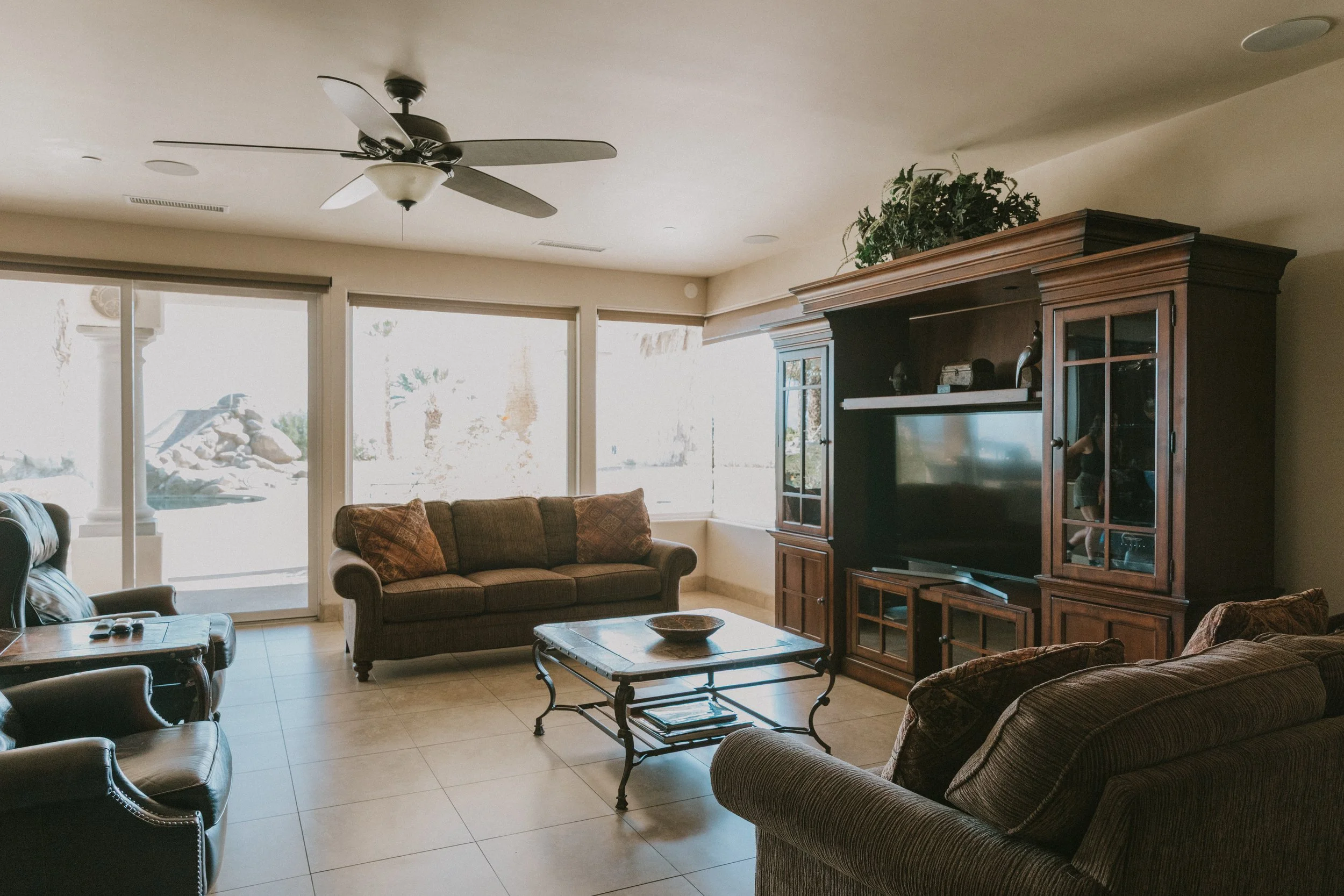 Living room with large windows, beige sofa, armchair, coffee table, entertainment center, and ceiling fan.