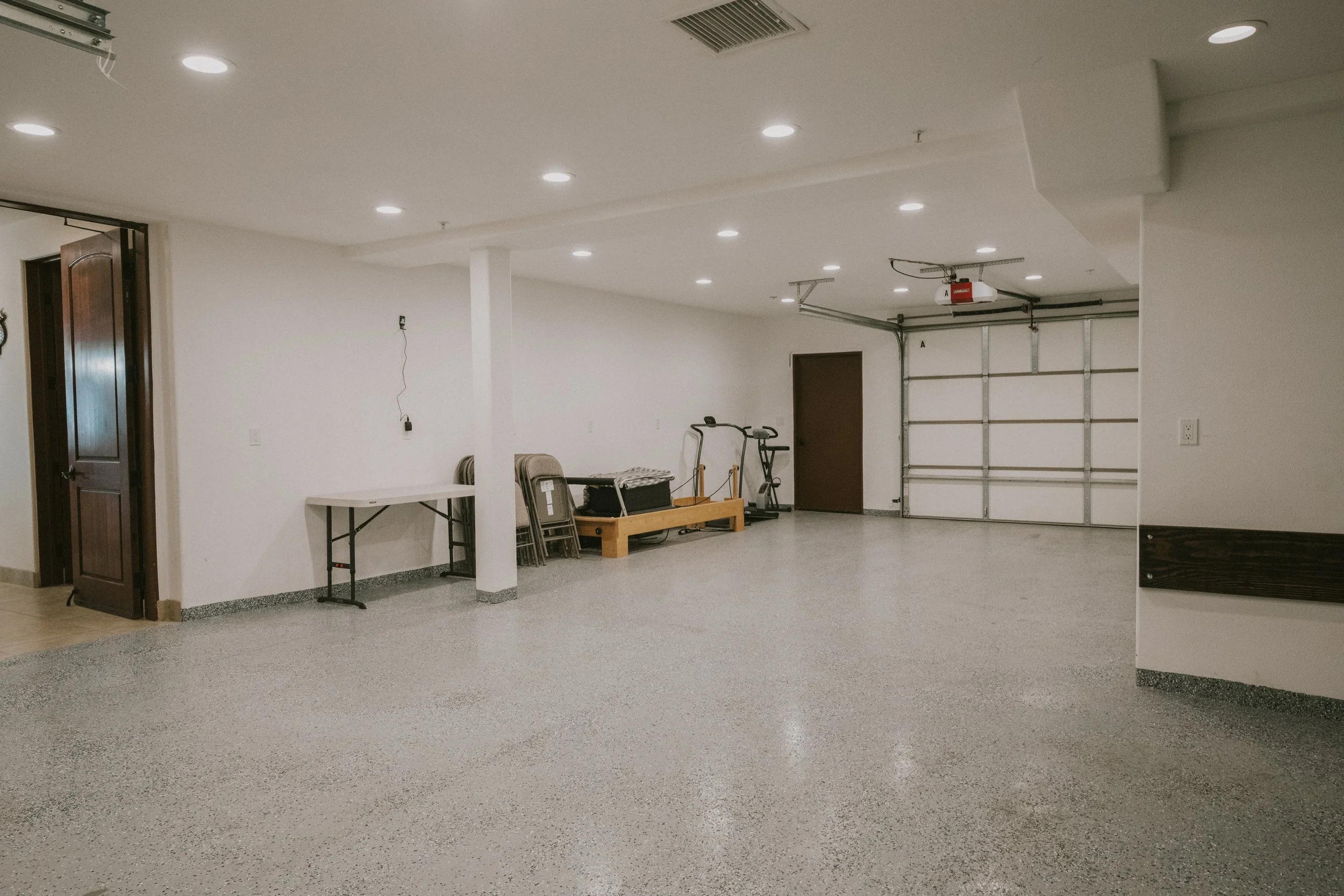 An empty garage with white walls, recessed ceiling lights, a partially open garage door, and various stored items including chairs, a treadmill, and a wooden cart.