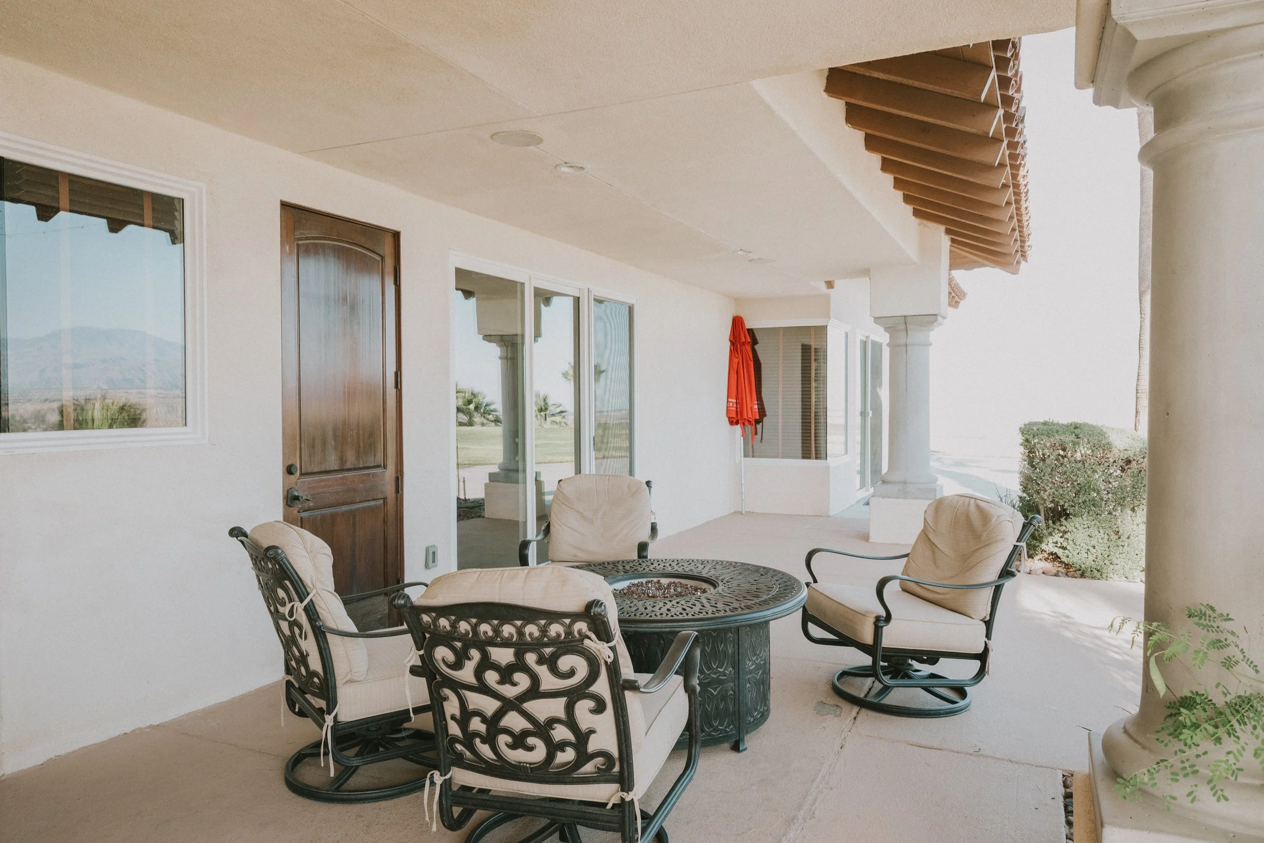 Covered patio area with outdoor furniture including a round metal table with ornate detailing and four cushioned chairs. A wooden door and sliding glass doors lead into the house. The ceiling has recessed lighting and decorative wooden beams. A red p