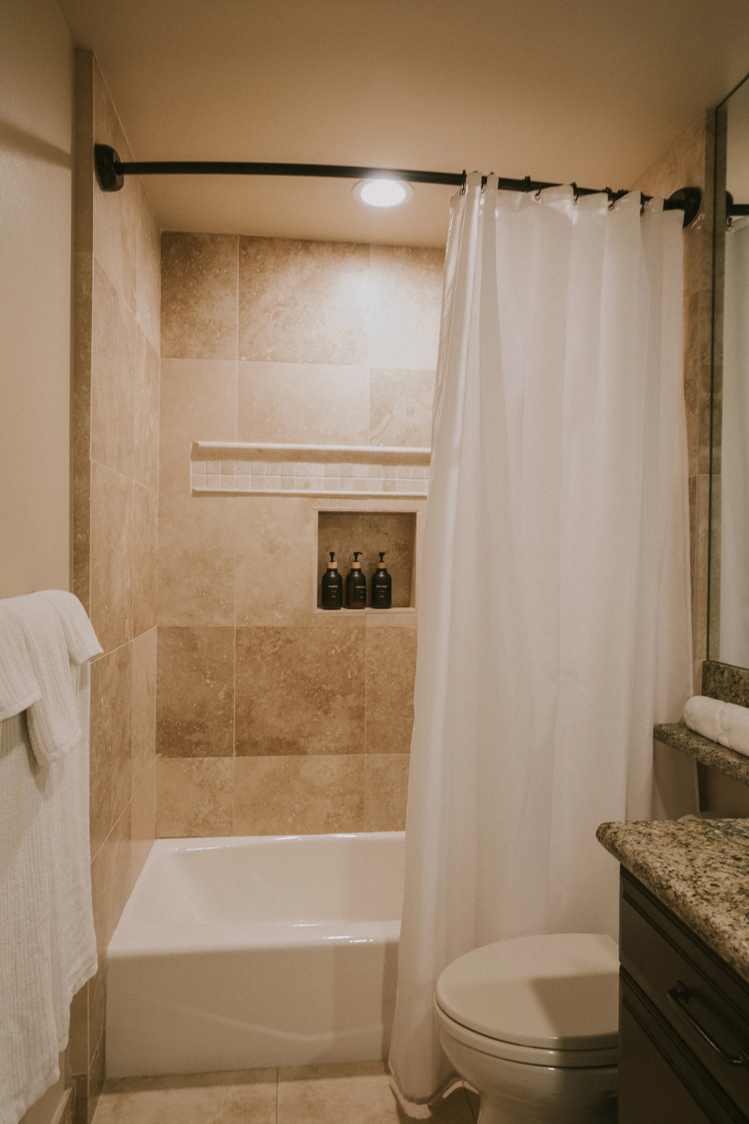 A bathroom with a shower area that has beige tiled walls and a built-in niche with shampoo bottles. There is a white shower curtain, a granite countertop, a mirror, and a white toilet. Towels are hanging on a towel rack.