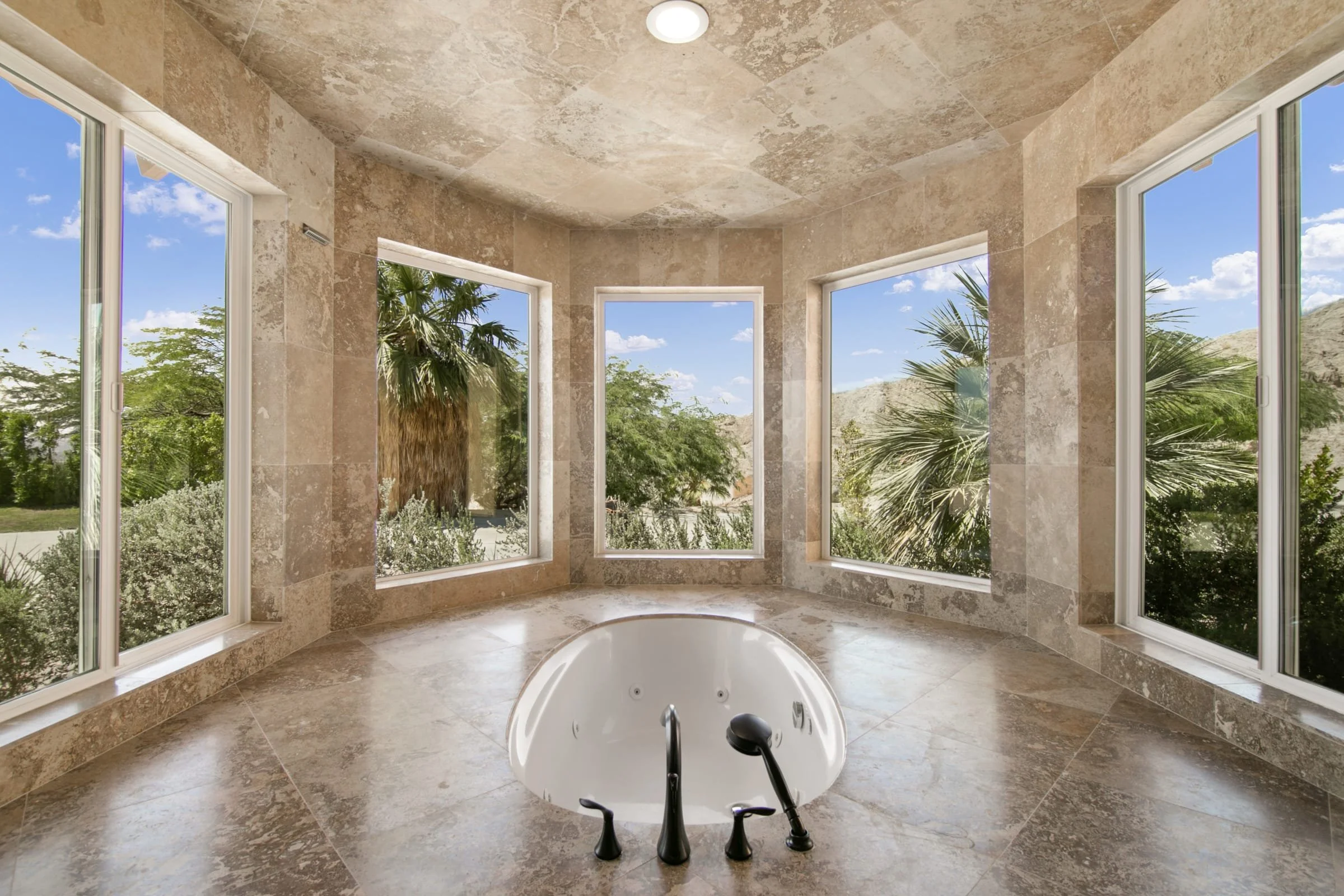 A luxurious bathroom with beige marble tiles and a corner bathtub with multiple jets, surrounded by large windows showing palm trees and a desert landscape.