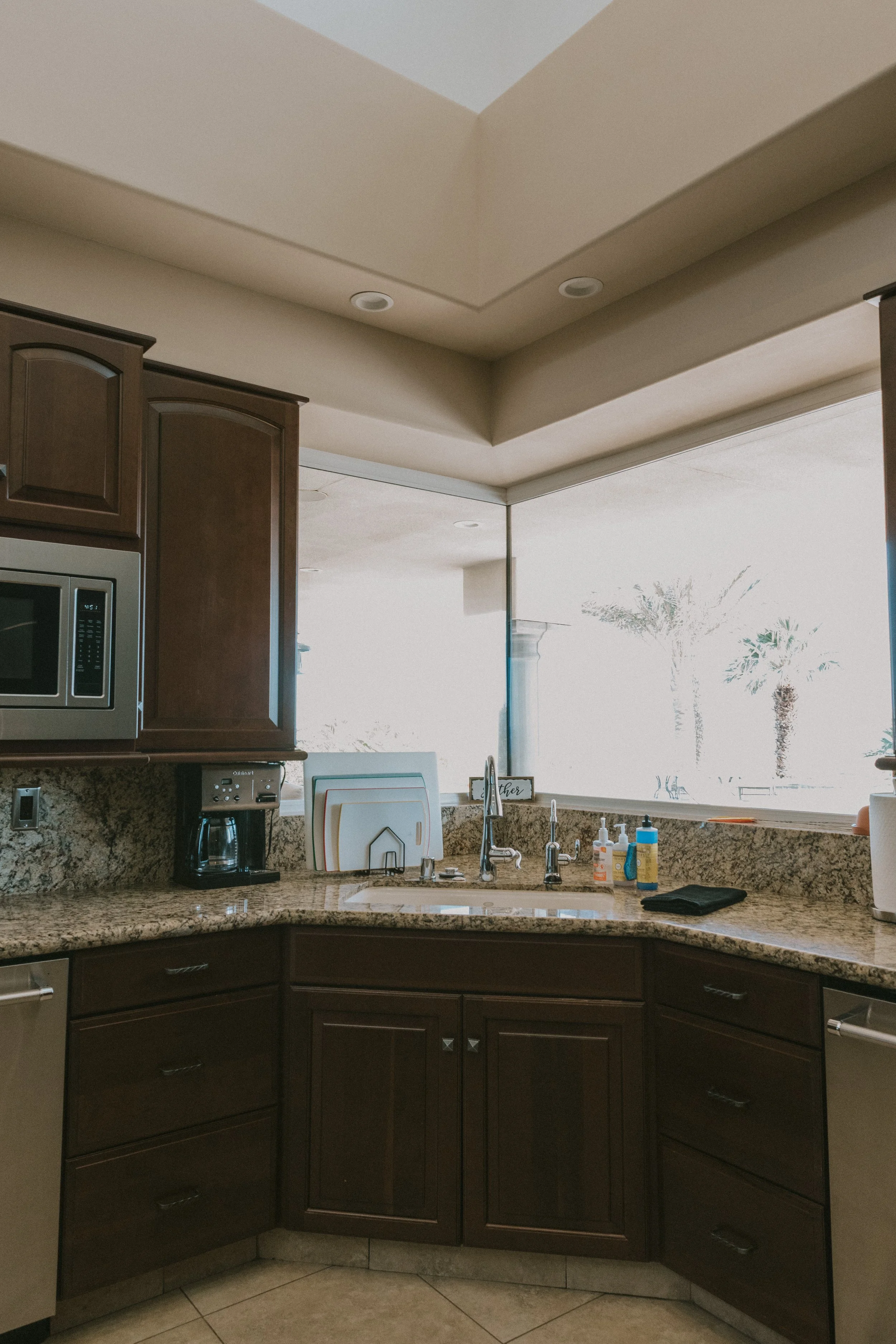 Kitchen with dark wood cabinets, granite countertop, stainless steel microwave, coffee maker, and a sink near a large window with a view of palm trees outside.