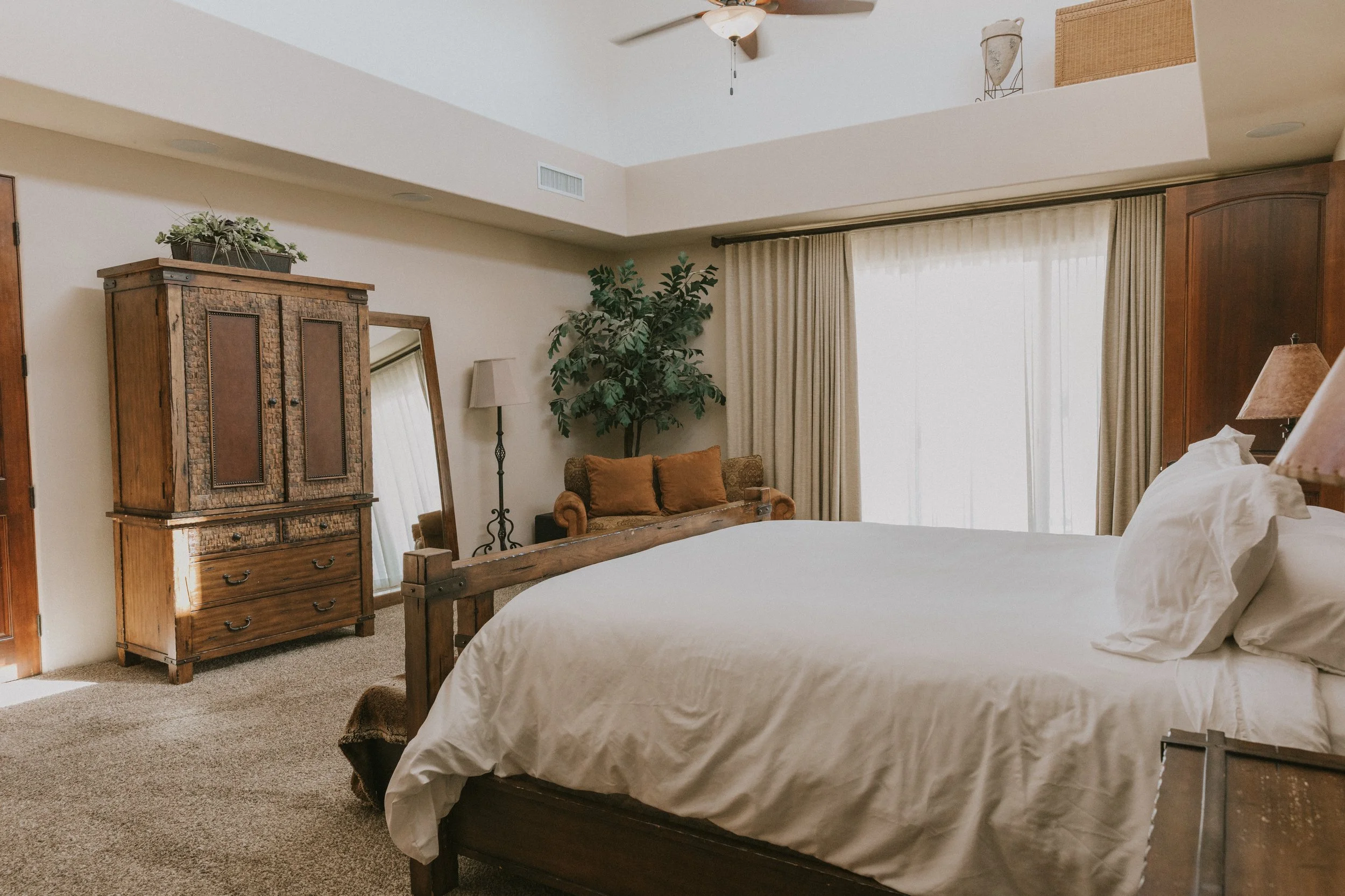 A cozy bedroom with a wooden bed frame, white bedding, a rustic wooden dresser, a mirror, a floor lamp, a large window with beige curtains, a potted plant, and a wicker basket on the upper balcony.