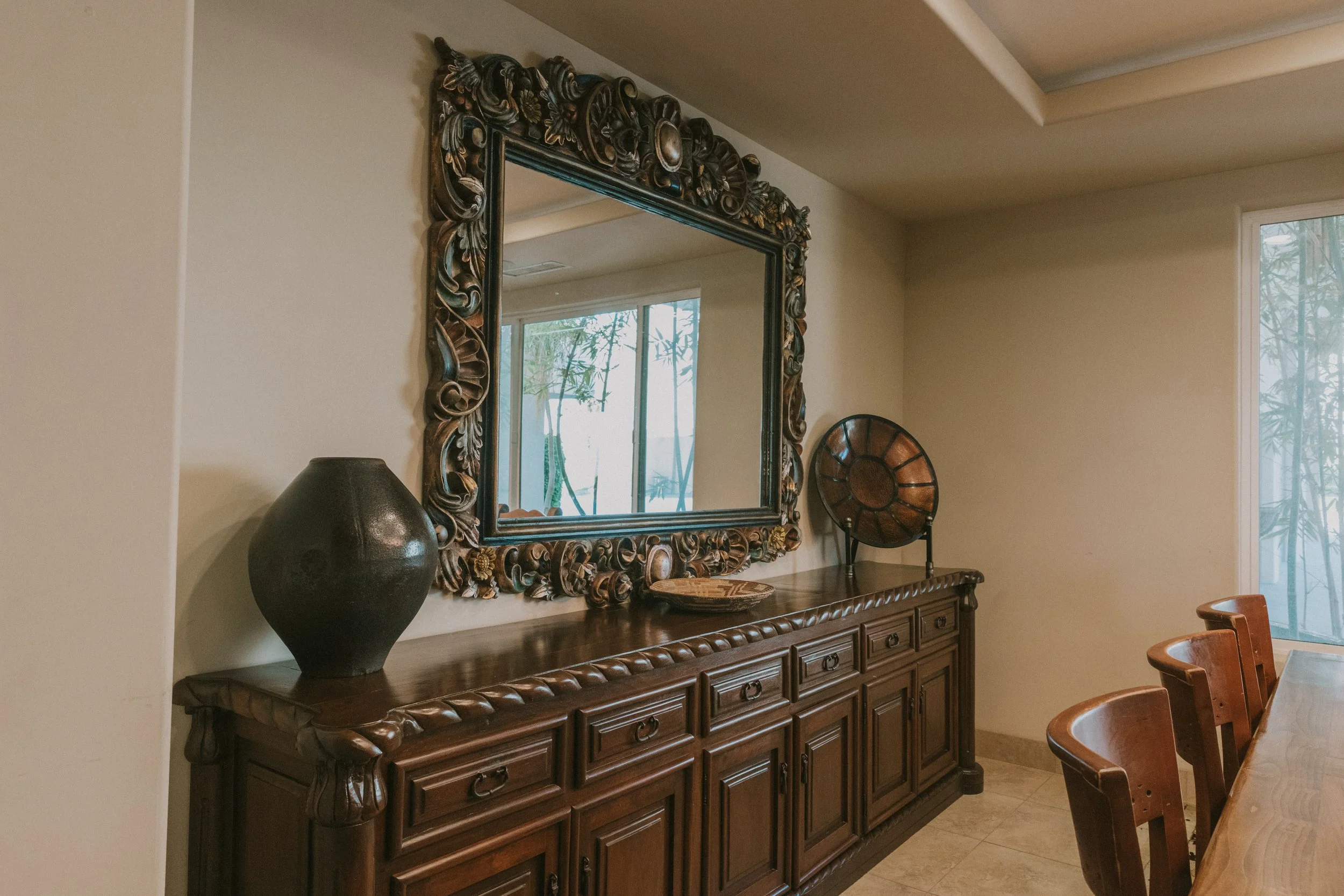 A wooden sideboard with drawers, topped by a large ornate mirror, a black vase, and a decorative woven bowl. A window shows green foliage outside, and part of a wooden chair is visible on the right.