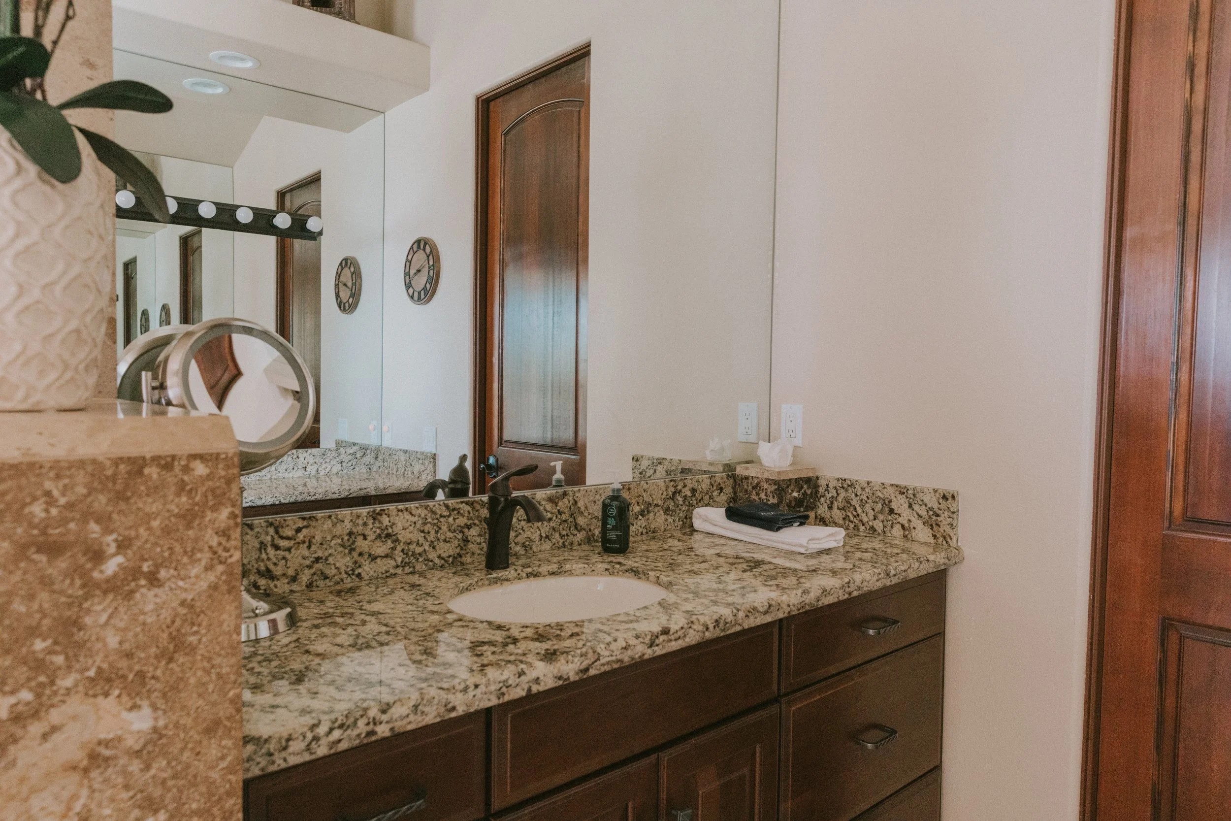 Bathroom vanity area with granite countertop, a sink with black faucet, soap dispenser, towel, and two electrical outlets, with a mirror, clock, and door reflected in the background.