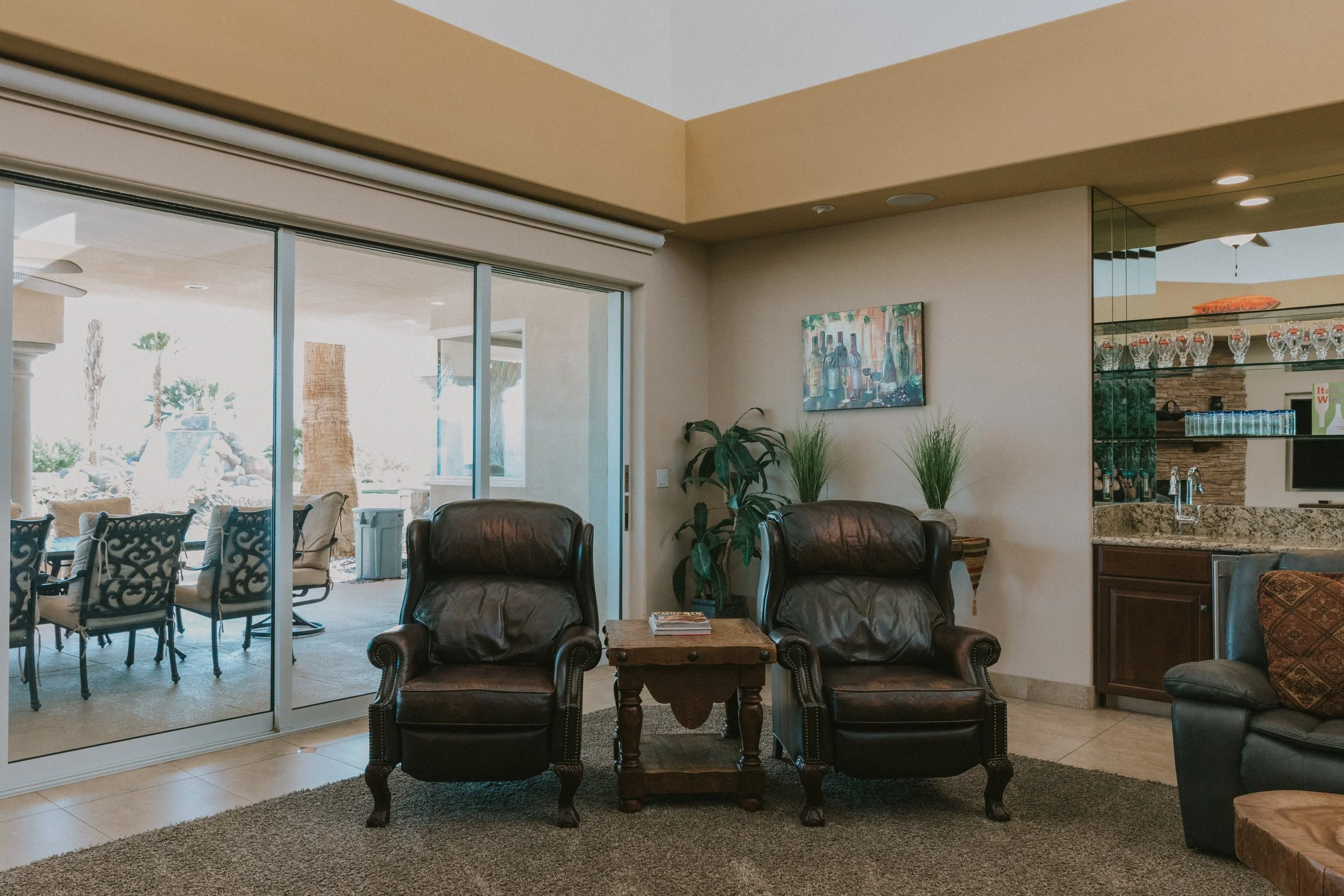 Living room area with leather chairs, a wooden table, houseplants, and a large sliding glass door leading to an outdoor patio with outdoor furniture and trees.