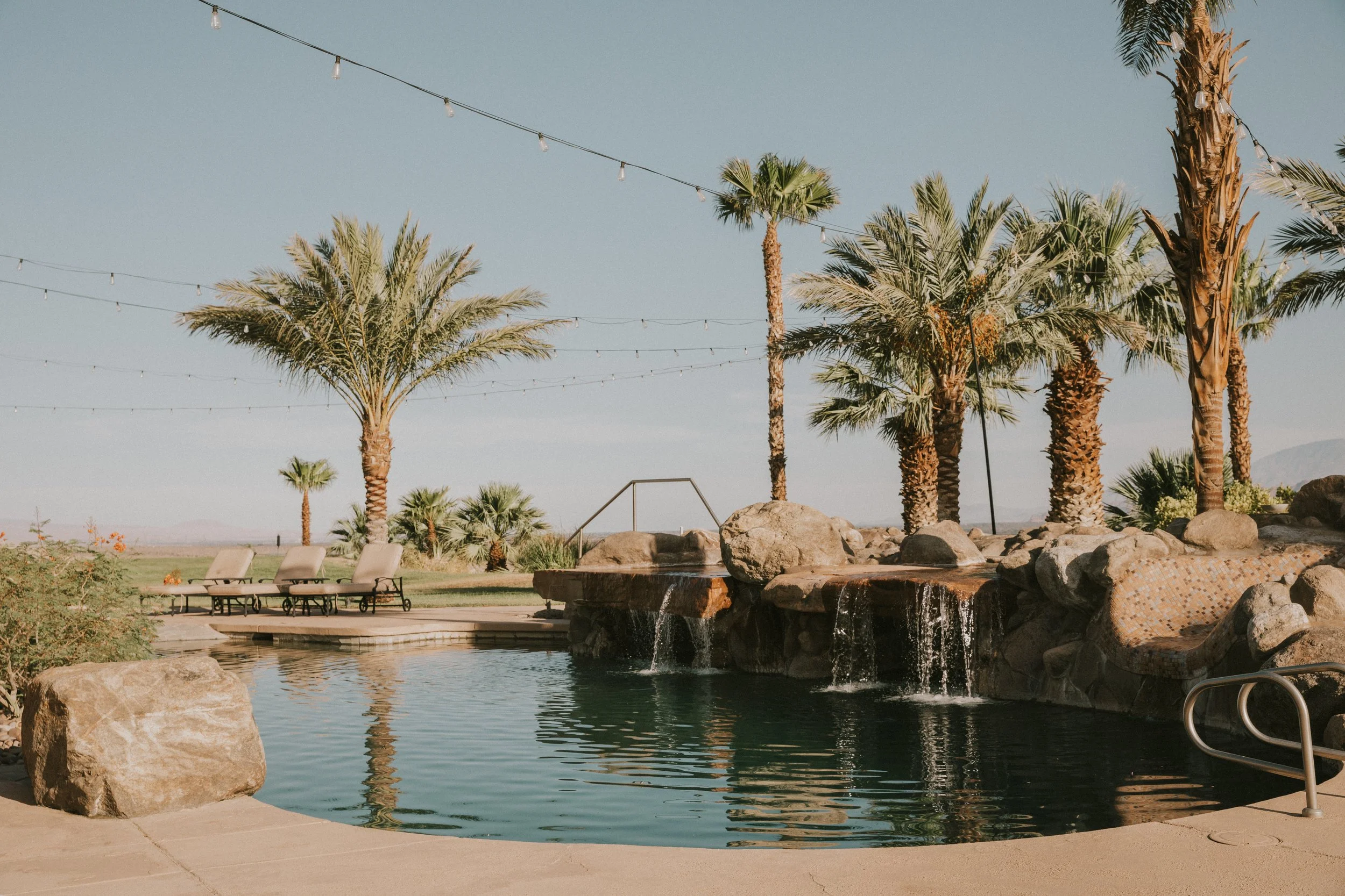 A swimming pool area with large rocks and a waterfall feature, palm trees, lounge chairs, and string lights above, set in a sunny outdoor environment.