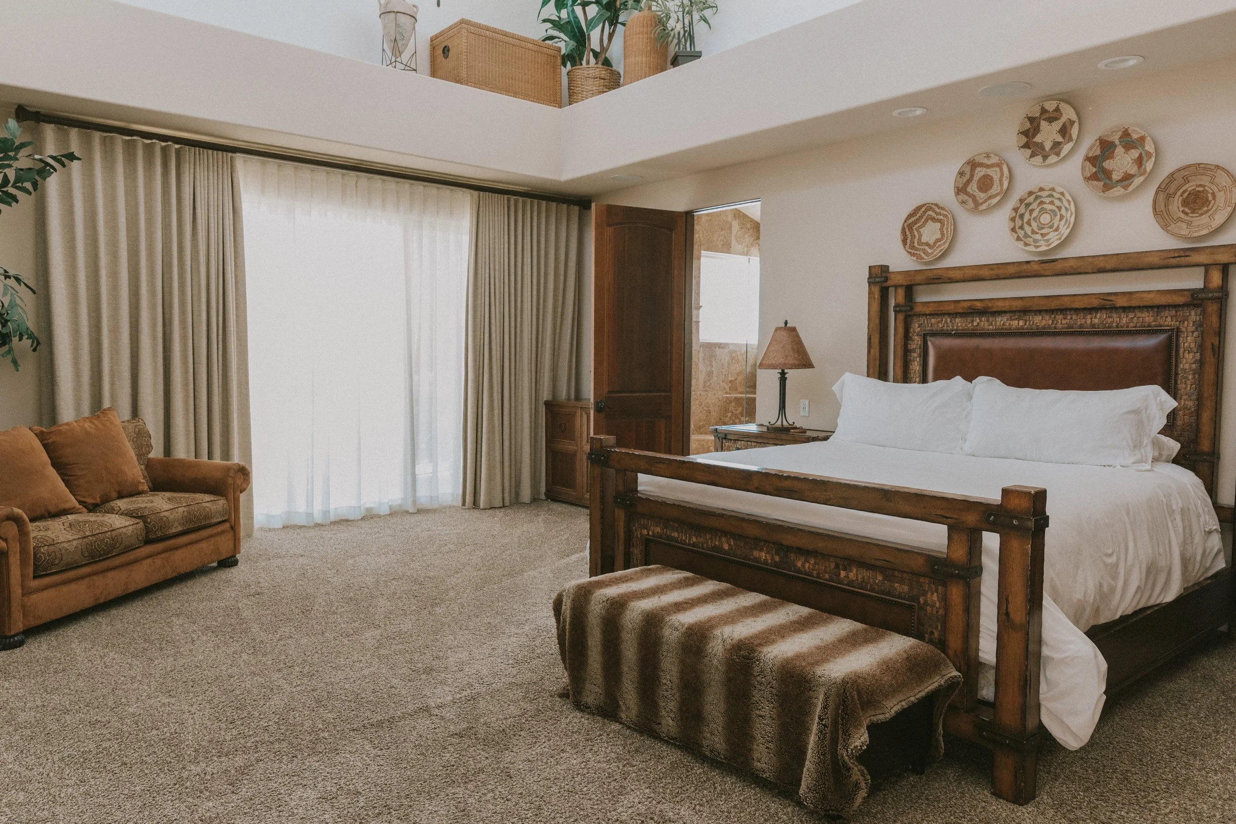 A cozy bedroom with a wooden bed frame, white bedding, a nightstand with a lamp, a brown patterned couch with pillows, large windows with curtains, and decorative plates on the wall.