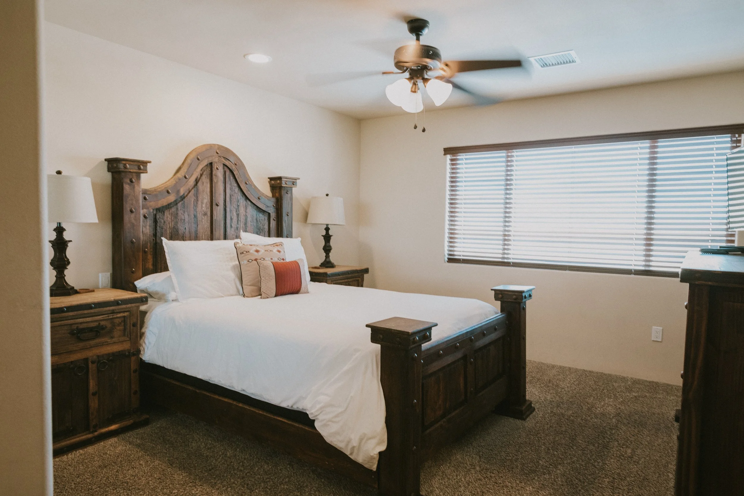 A bedroom with a wooden bed frame, white bedding, pillows, two matching bedside tables with lamps, a window with horizontal blinds, a ceiling fan with light, and carpeted floors.