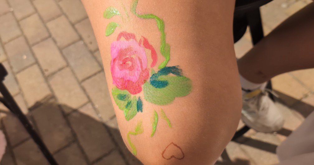A henna tattoo of a pink flower with green leaves on a person's forearm, with a small heart outline below it. The background shows a paved surface and part of a person's leg and shoe.