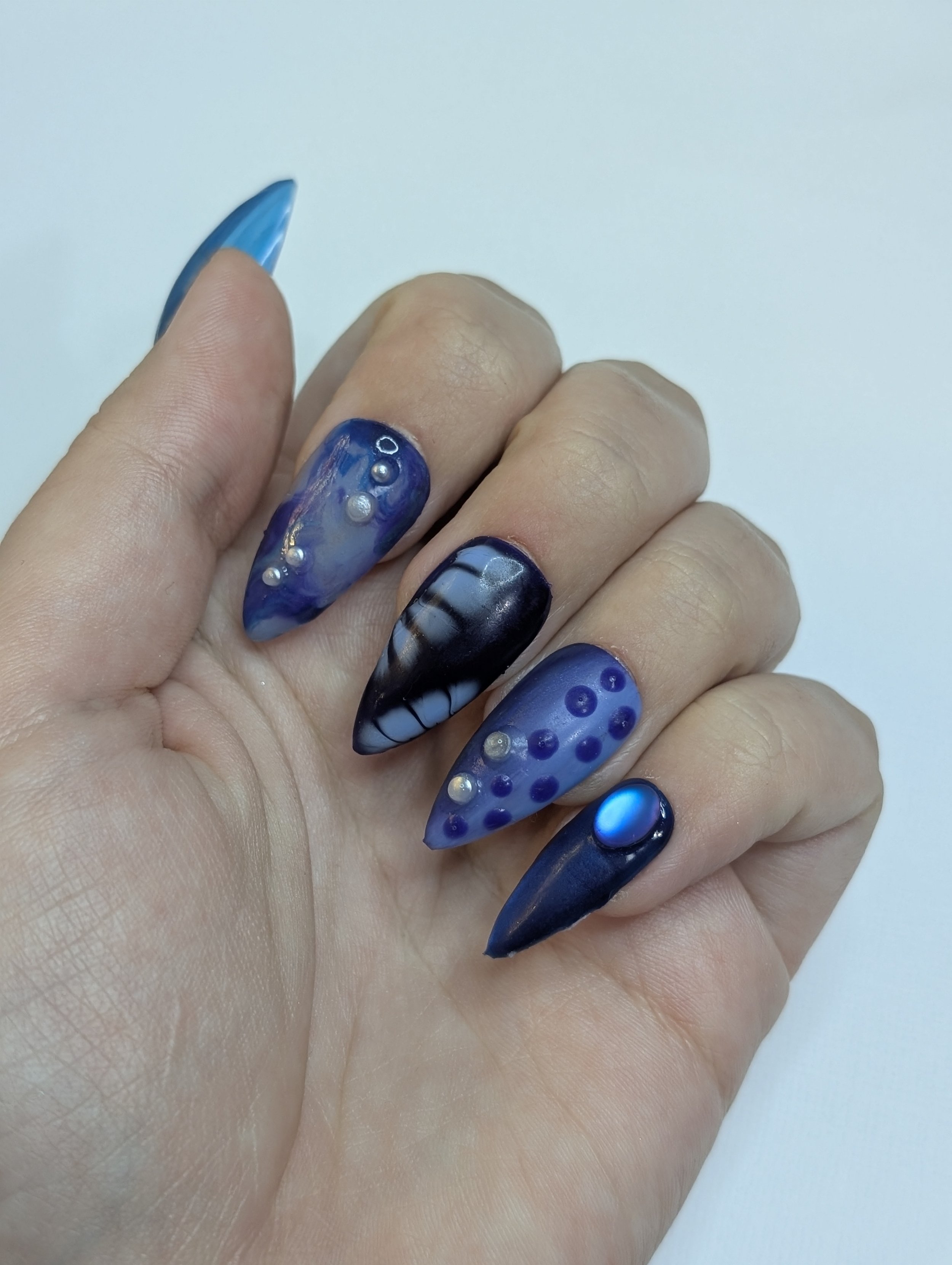 Close-up of a hand with long, decorated blue and black gel nails, some adorned with small pearl-like embellishments and intricate patterns.