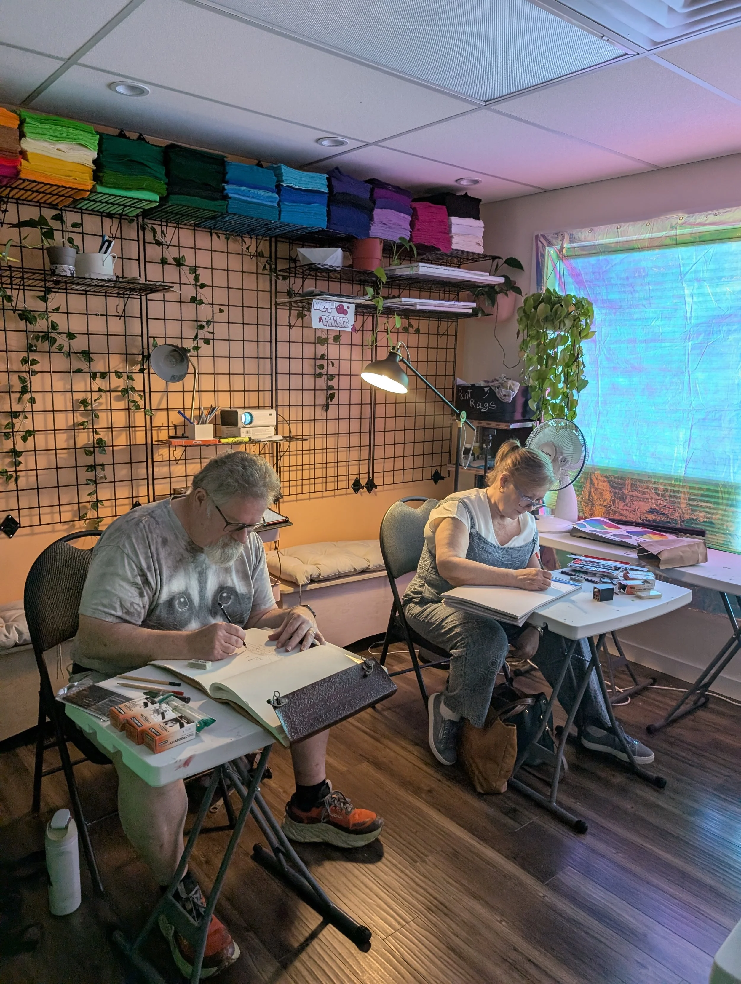Two people sit at a table working on art projects in a room decorated with colorful fabric and plants, with natural and artificial lighting.