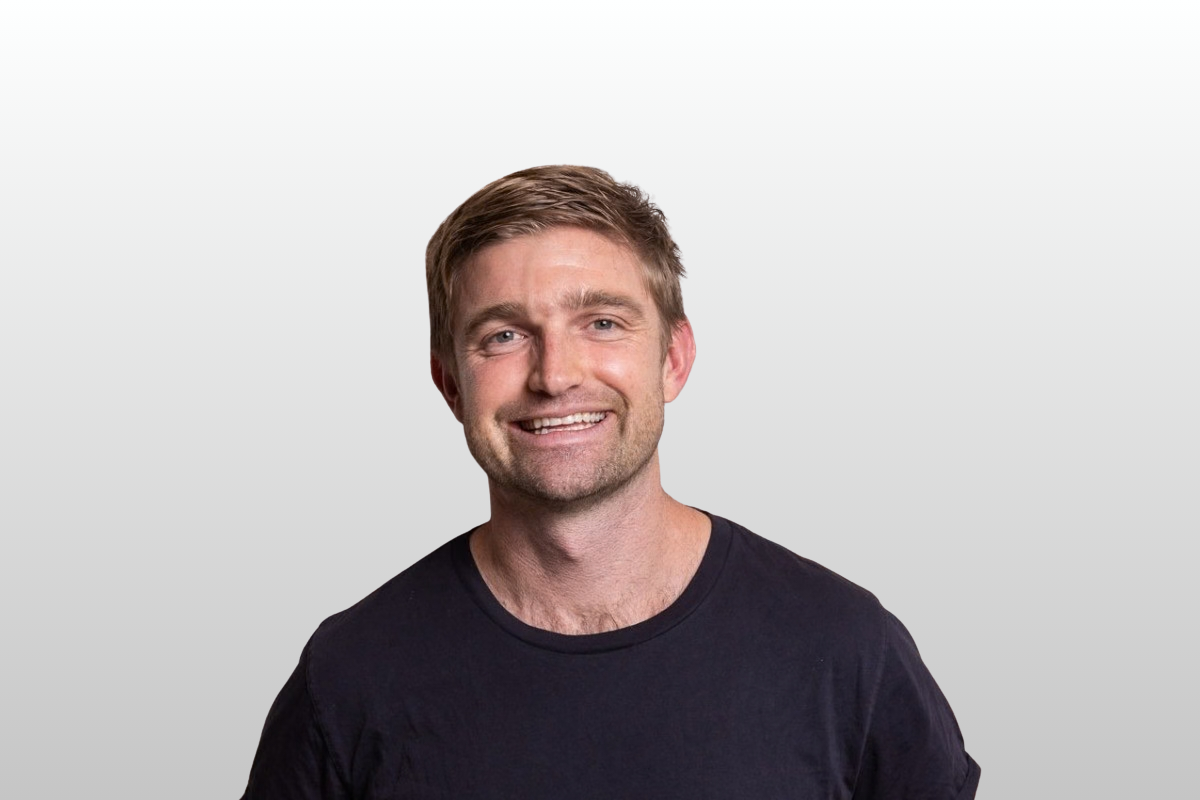 A smiling man with short light brown hair wearing a black t-shirt against a light gray background.