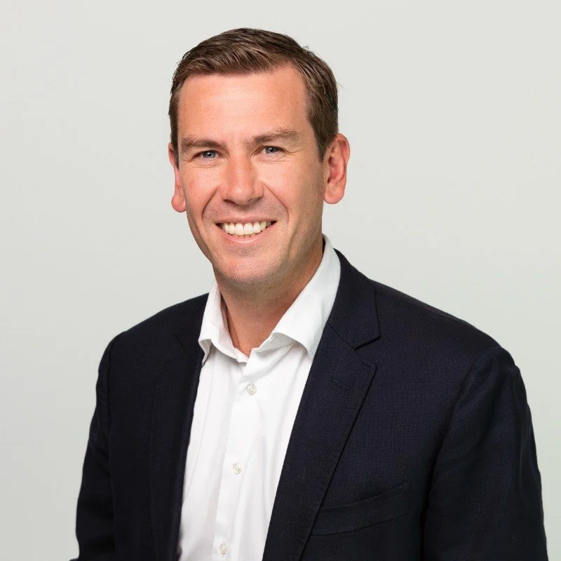 Professional headshot of a man with short brown hair, blue eyes, smiling, wearing a white shirt and dark blazer, against a plain light background.