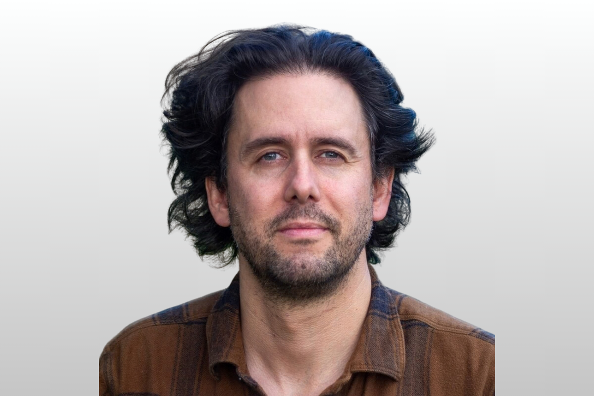 A man with dark, wavy hair and a light beard, wearing a brown shirt, stands against a neutral background.