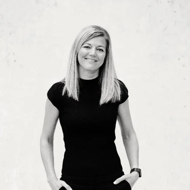Smiling woman with straight blonde hair, wearing a black short-sleeve top and a watch on her left wrist, standing against a plain light background.