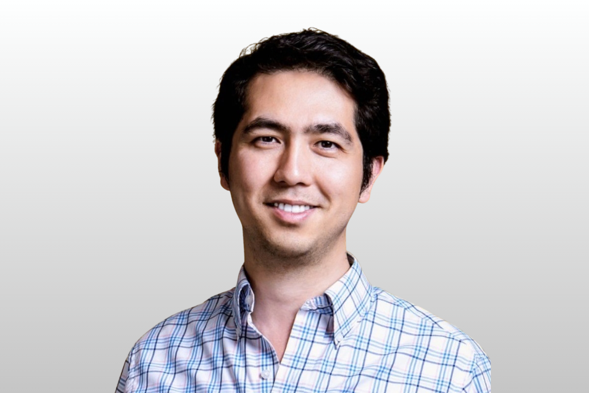 A smiling man with dark hair wearing a checked shirt poses in front of a plain light-colored background.