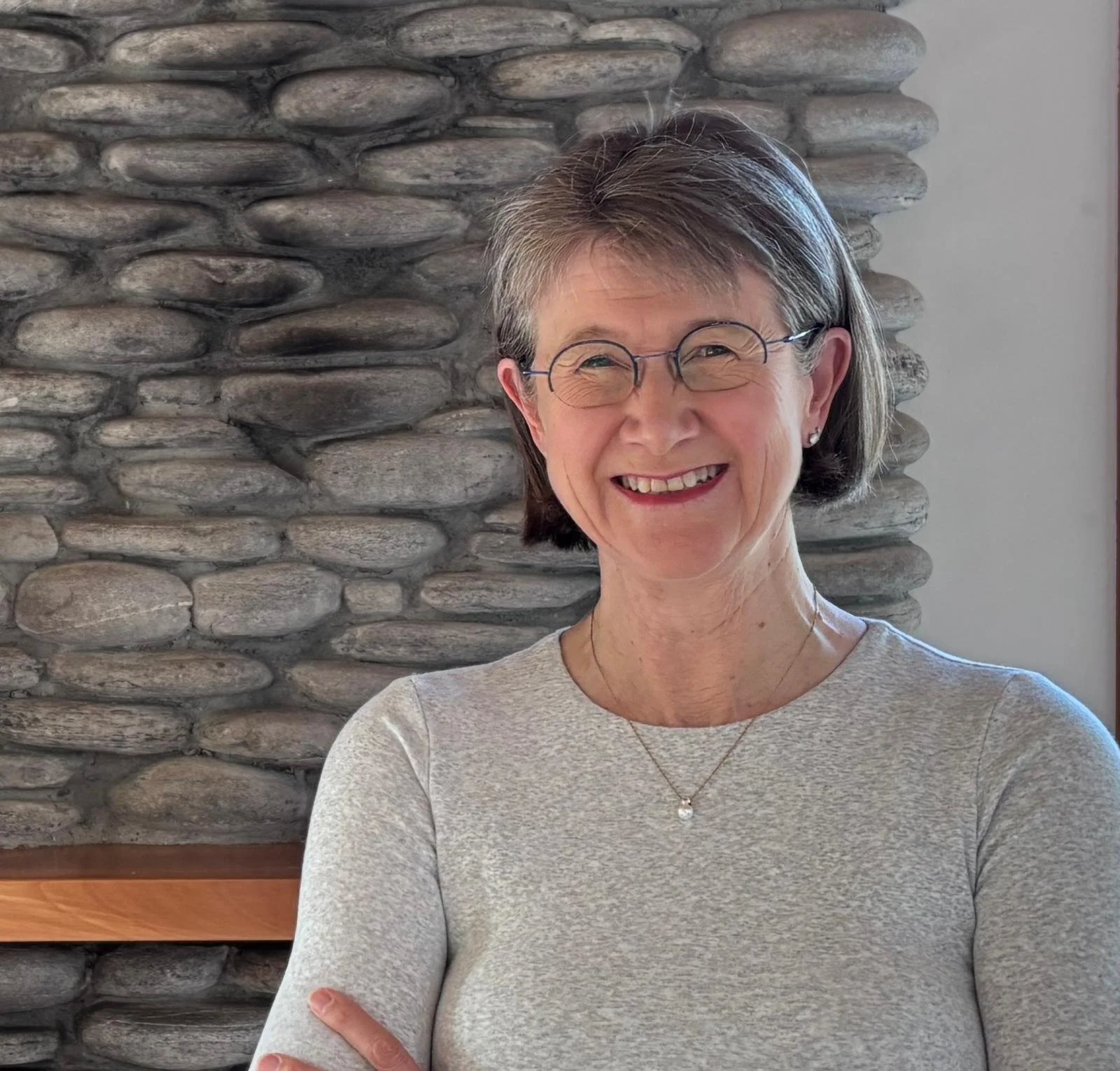 A smiling older woman with short gray hair and glasses, wearing a light gray top, standing in front of a stone wall.
