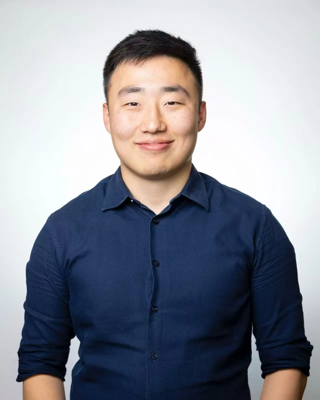 Portrait of a young man with short black hair smiling, wearing a navy blue dress shirt against a plain white background.