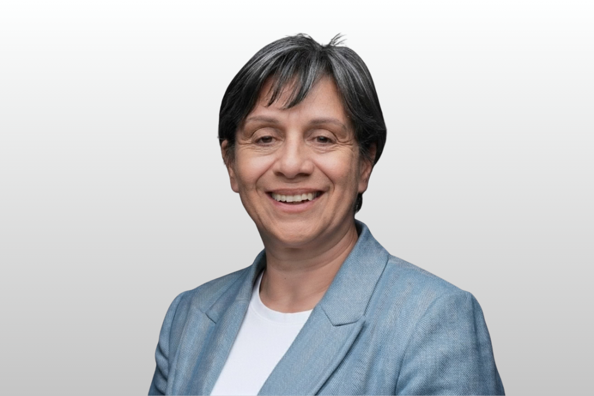 A woman with short gray hair smiling, wearing a light blue blazer over a white shirt, standing against a light gray background.