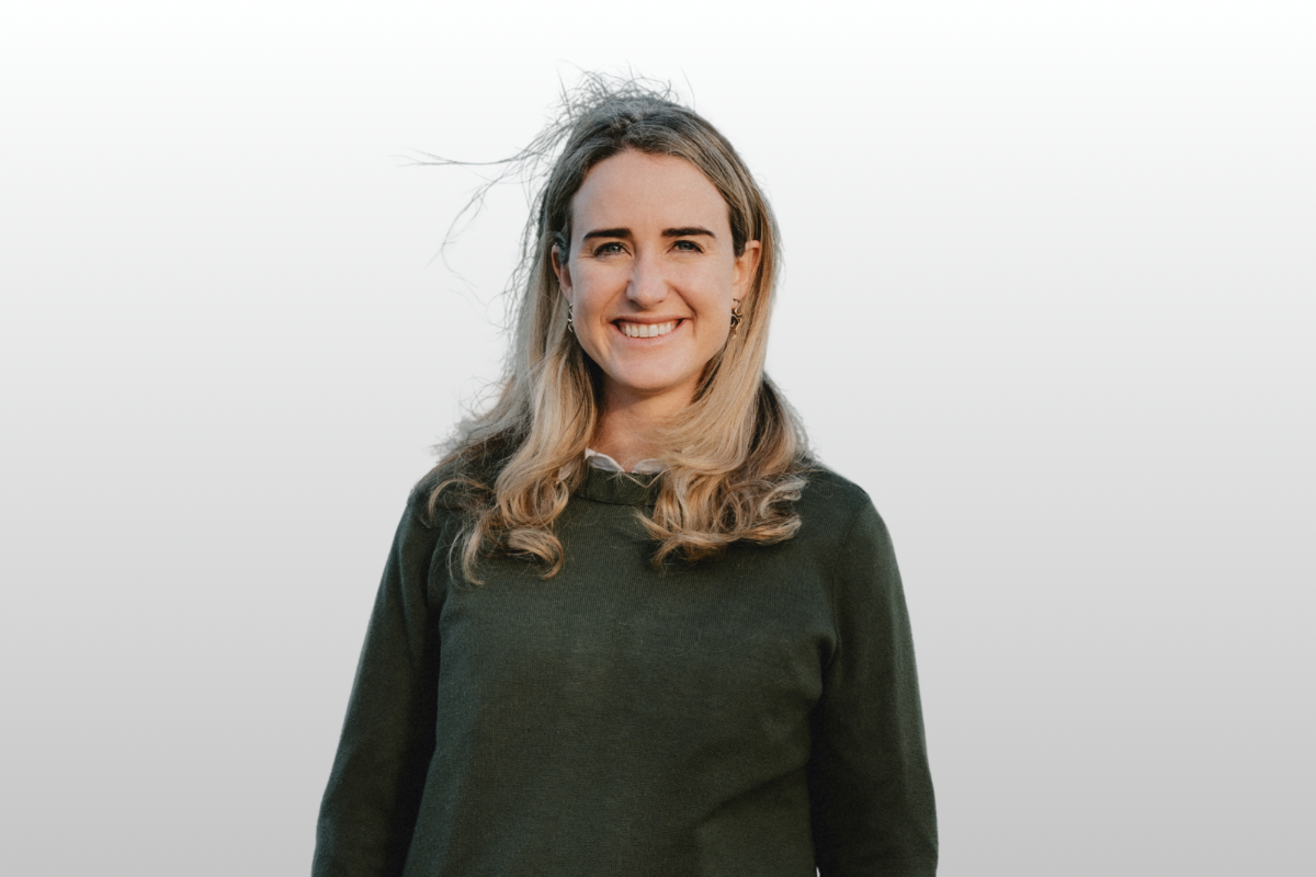 A woman with long, wavy blonde hair smiling outdoors with wind blowing her hair, against a plain background.