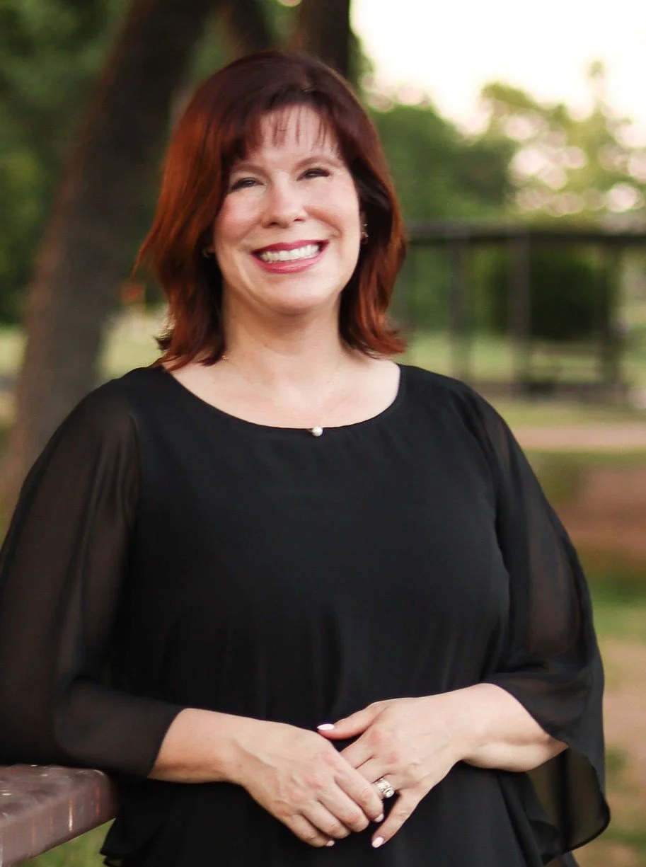 A smiling woman with shoulder-length reddish-brown hair, wearing a black dress with semi-sheer sleeves, standing outdoors next to a wooden surface, with a green, blurred background of trees and sky.