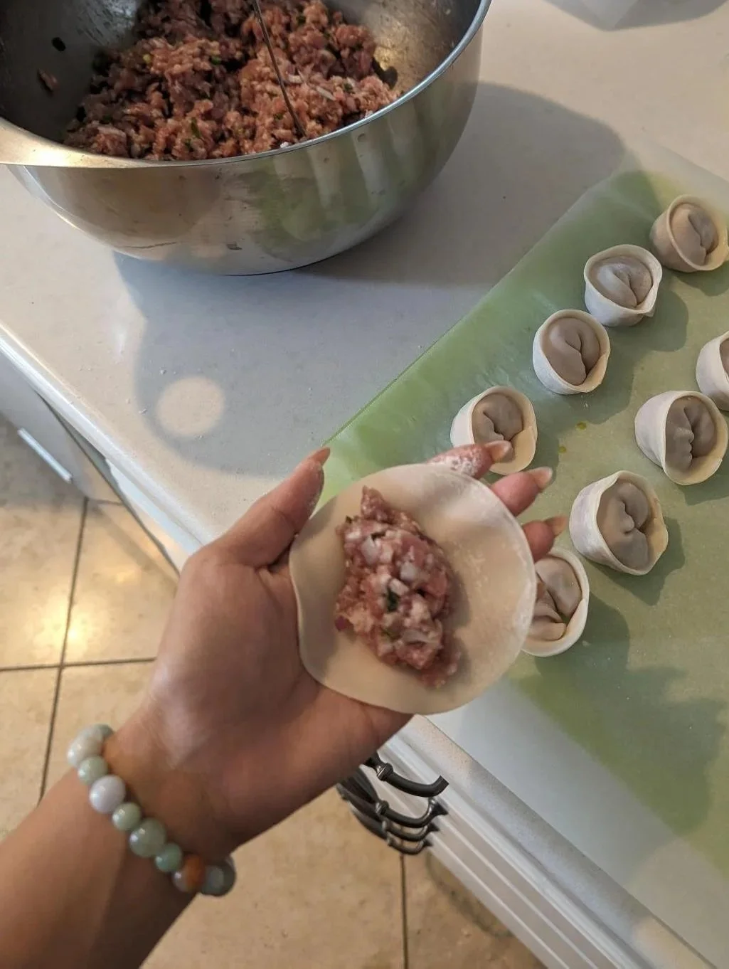 Woman preparing dumplings with ground meat filling, placing it inside round dough wrappers on a green cutting board.