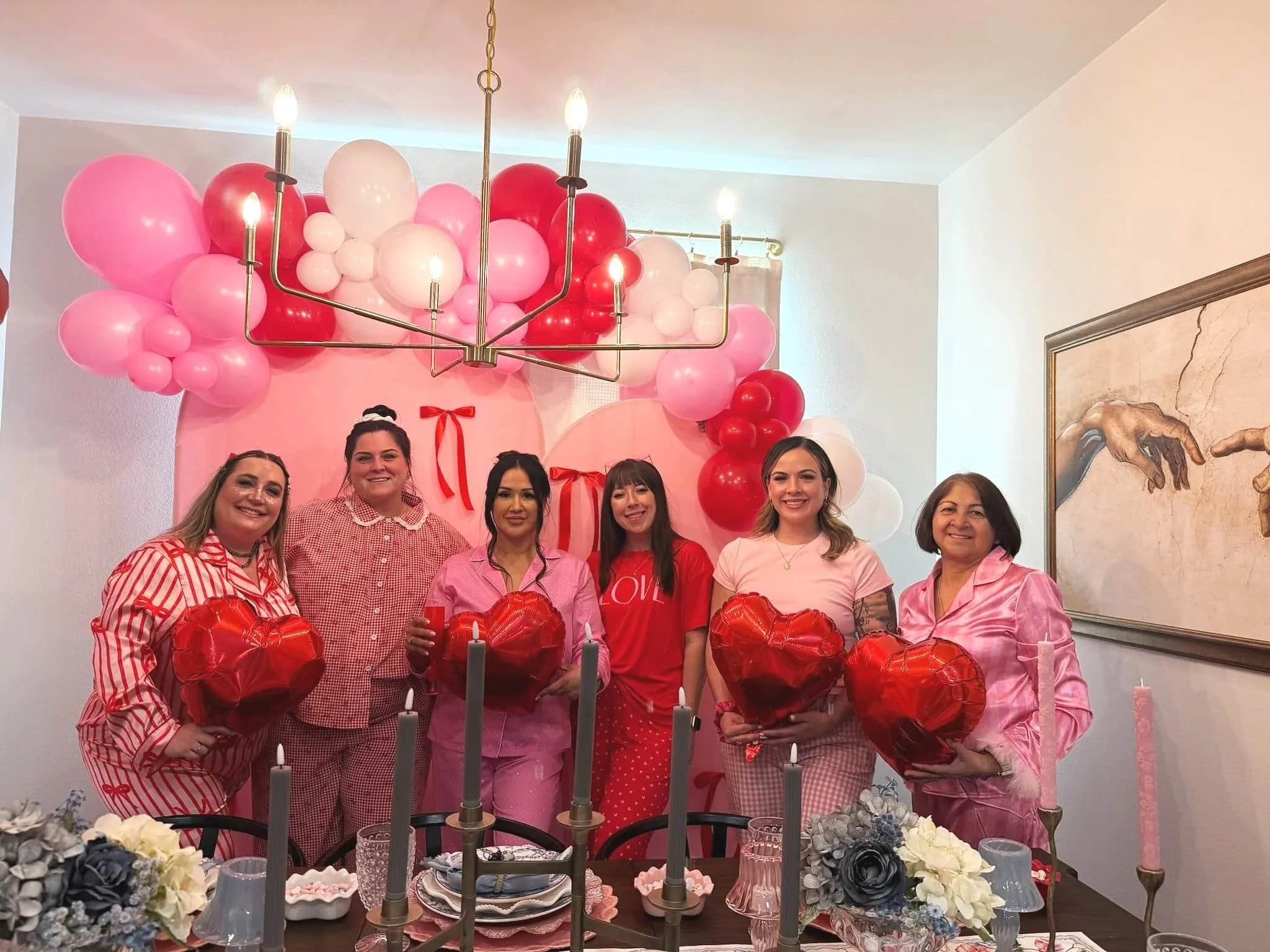 Six women in pink and red pajamas hold heart balloons at a table with flowers, celebrating in a room decorated with balloons.